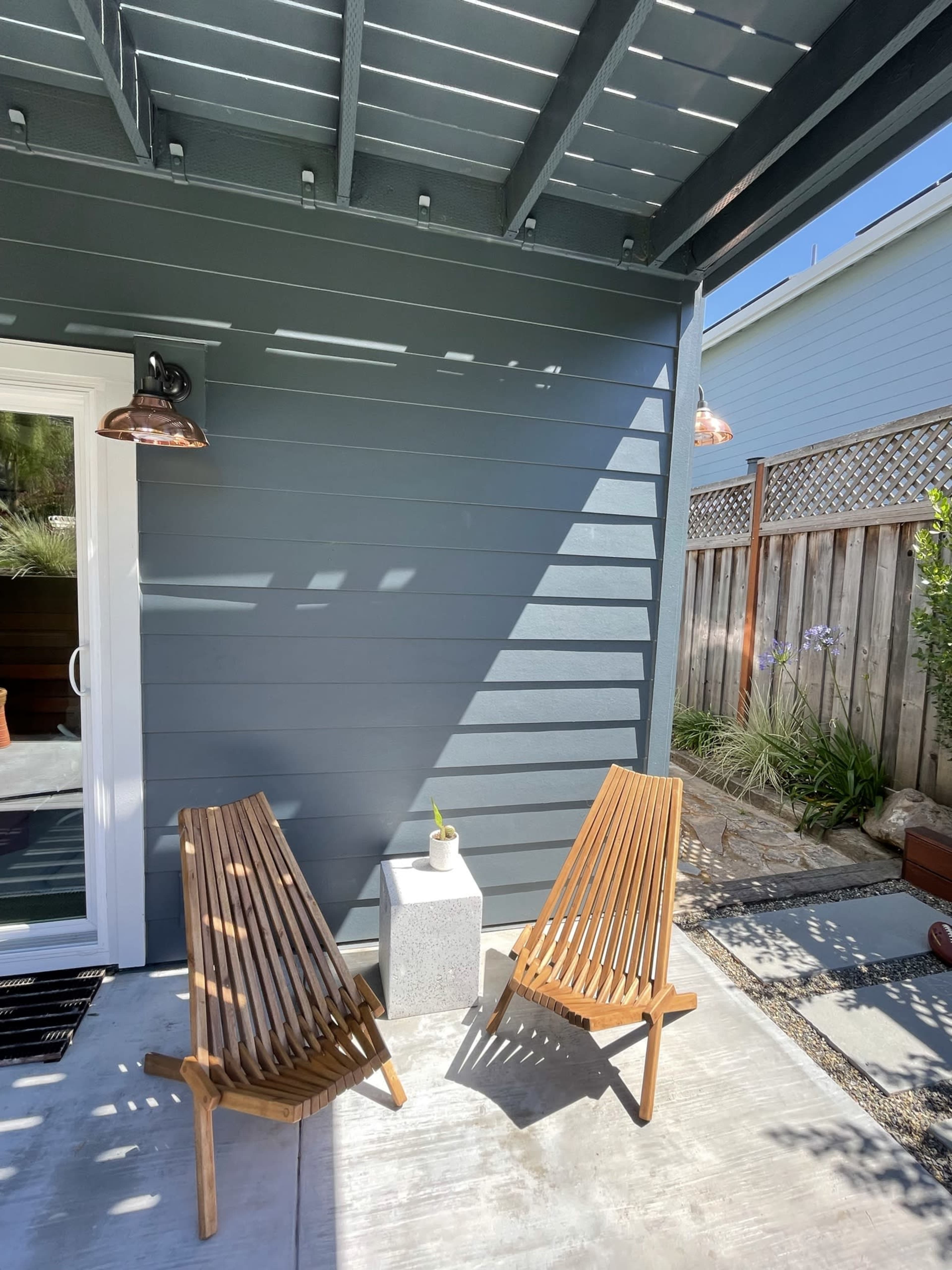 Two wooden lounge chairs beside a small concrete table on a concrete patio next to a gray house wall.
