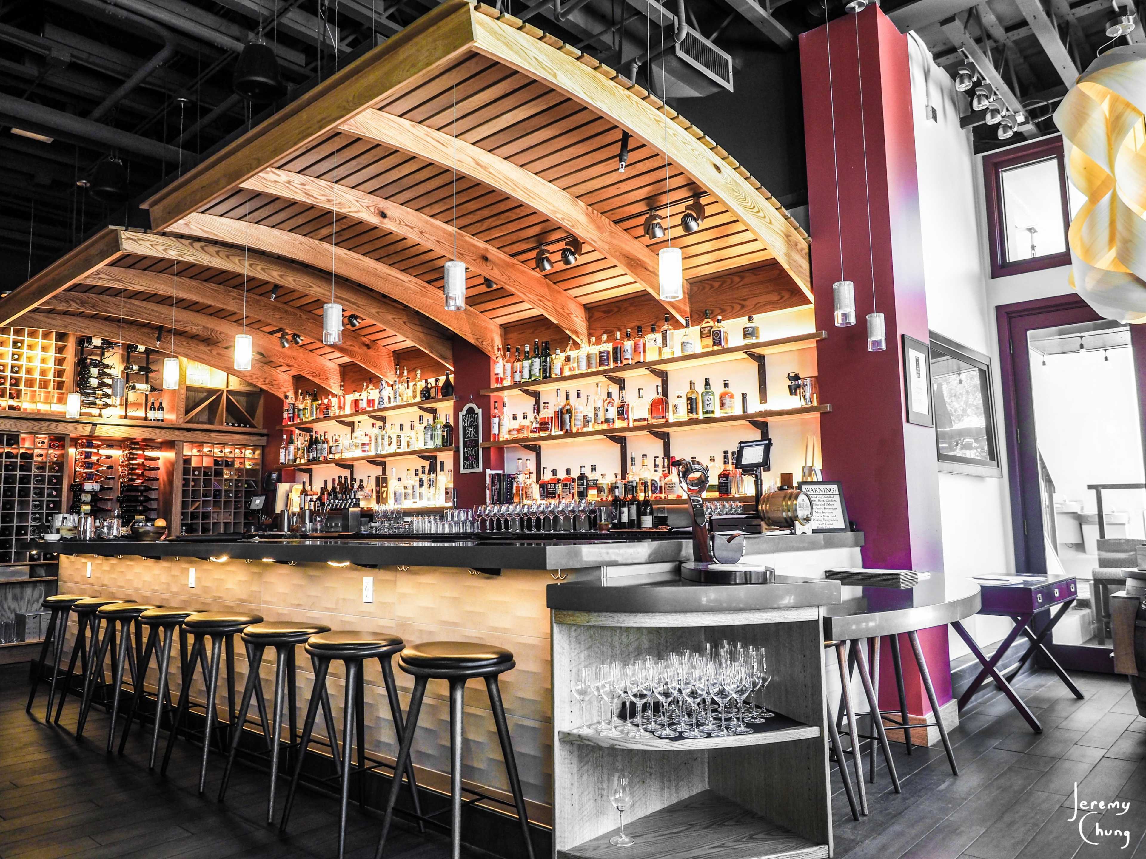 A modern bar with a curved wooden ceiling, bottles displayed on shelves in the background, and a sleek countertop with stools.