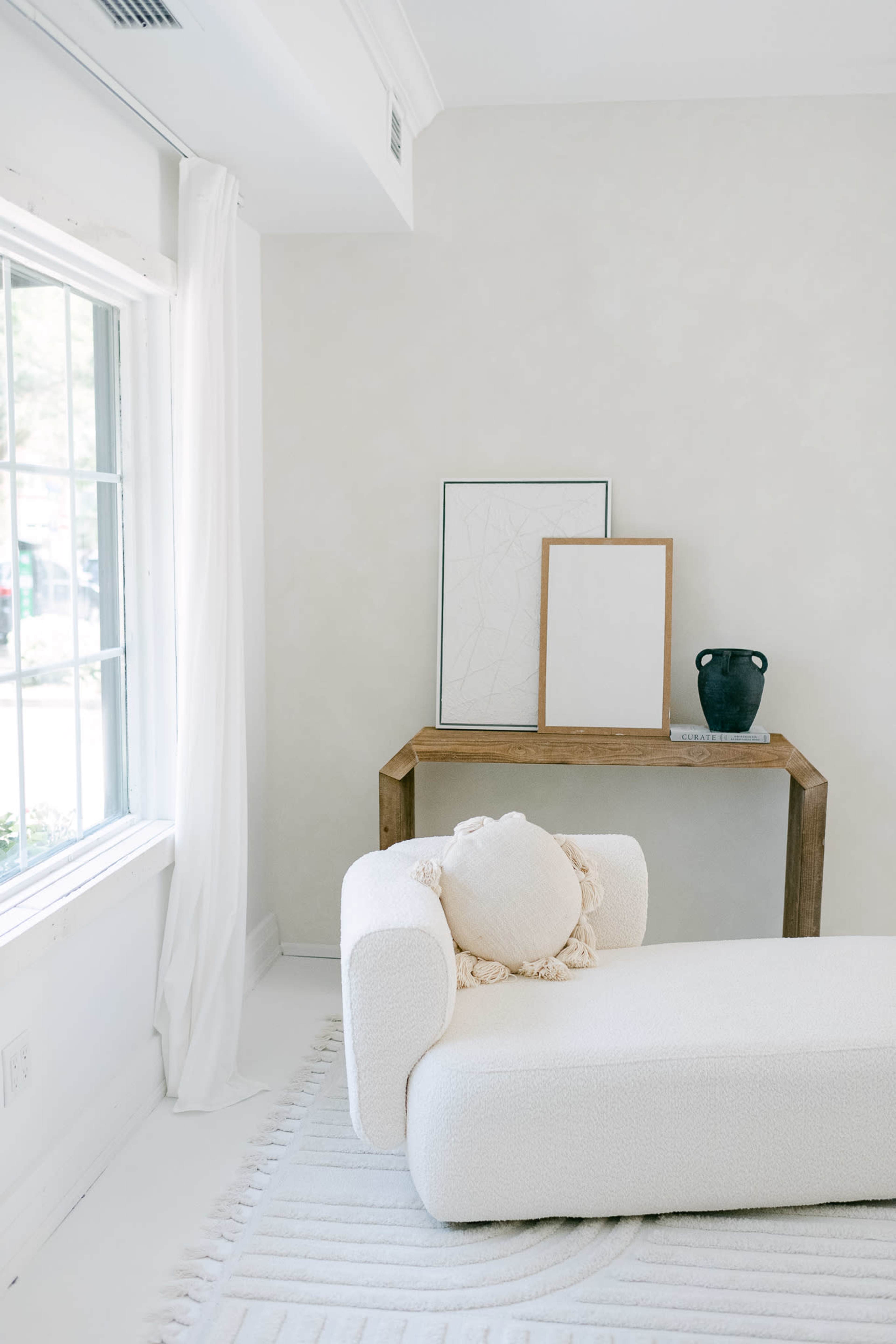 A minimalist living space with a white sofa, a wooden table displaying two empty frames and a vase, and a large window letting in natural light.