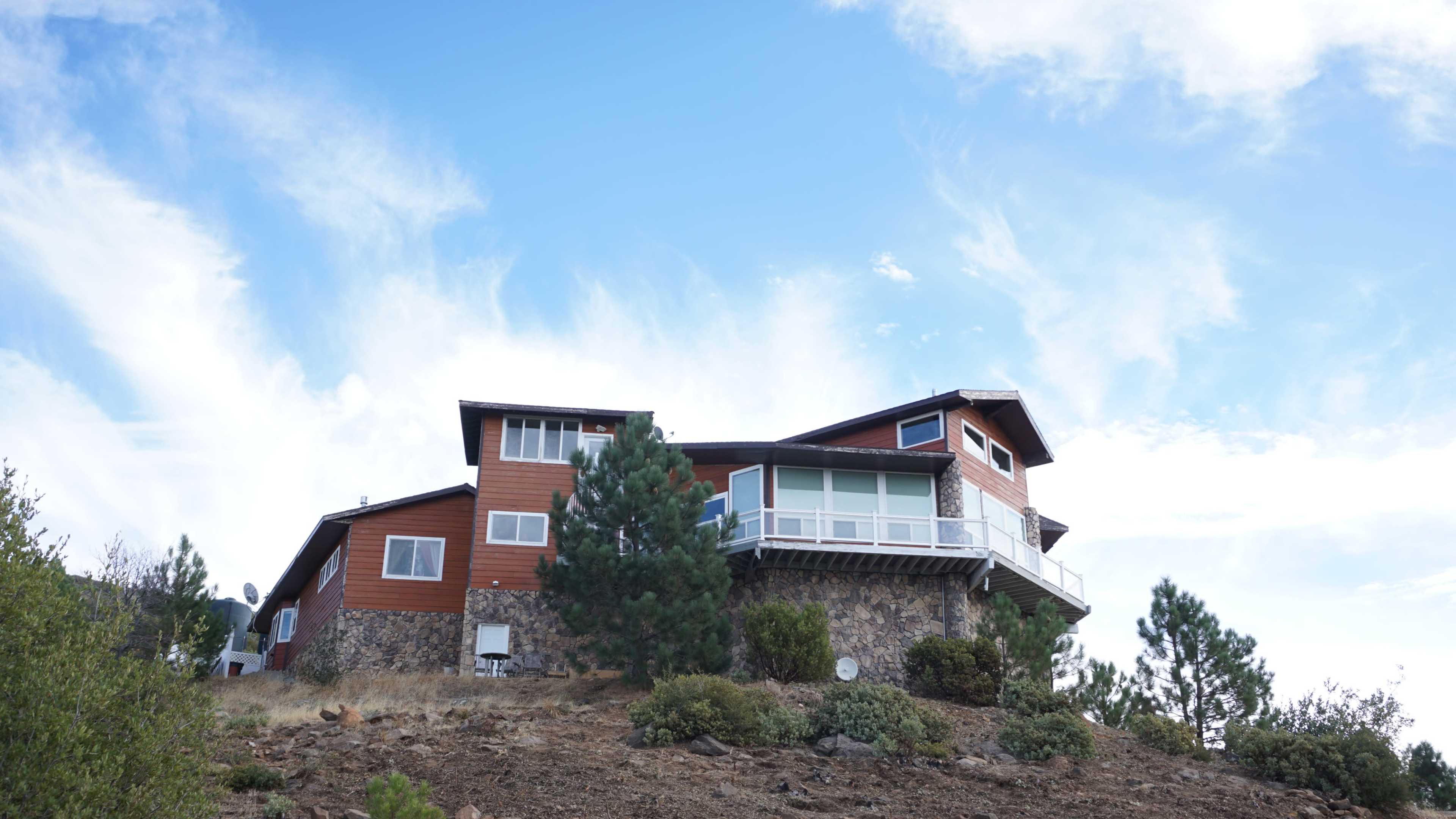 A large, multi-level house with a stone foundation is situated on a hillside, surrounded by pine trees and rocky terrain under a partly cloudy sky.