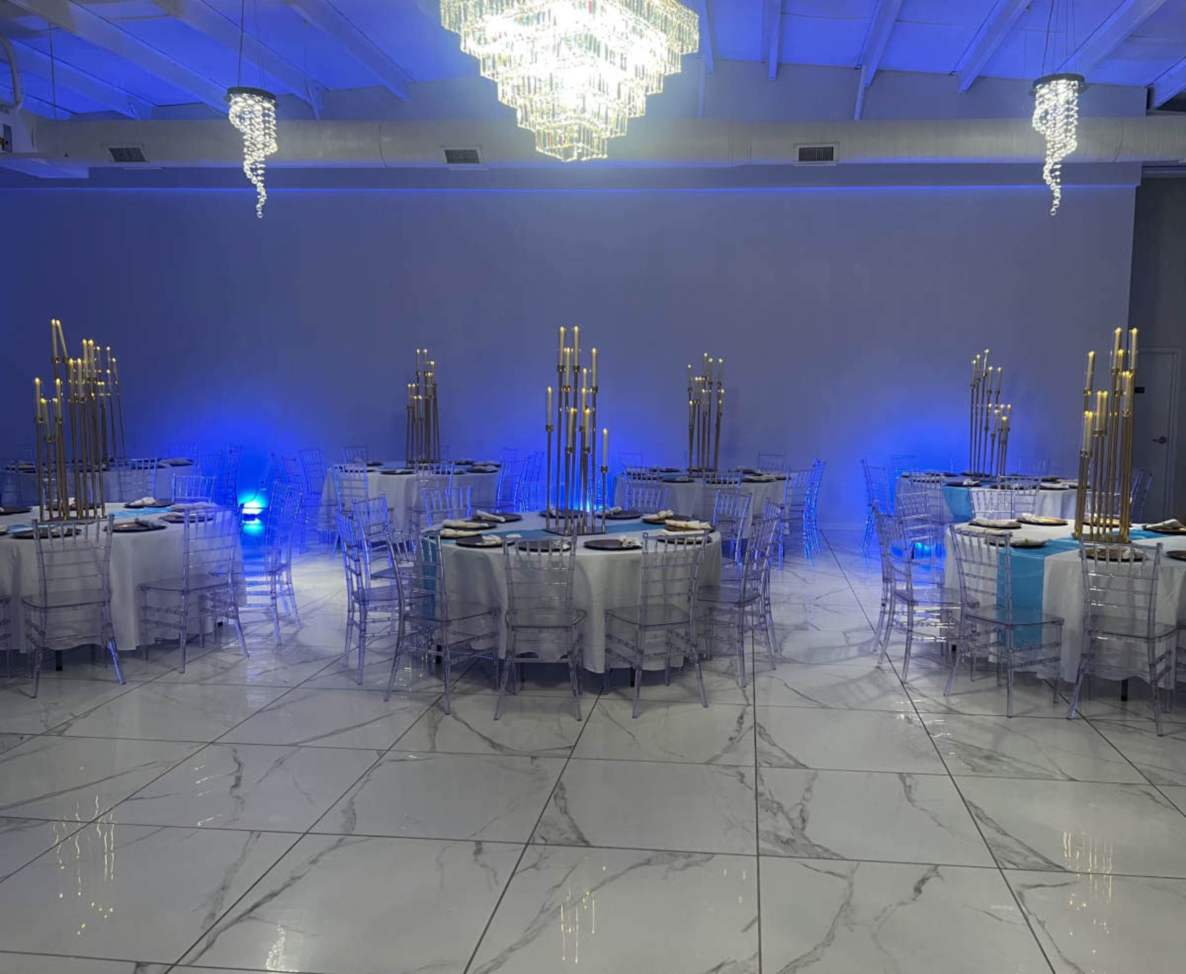 The image shows an elegant banquet hall set up with round tables, each adorned with tall golden candleholders, against a backdrop of blue lighting and a large chandelier.