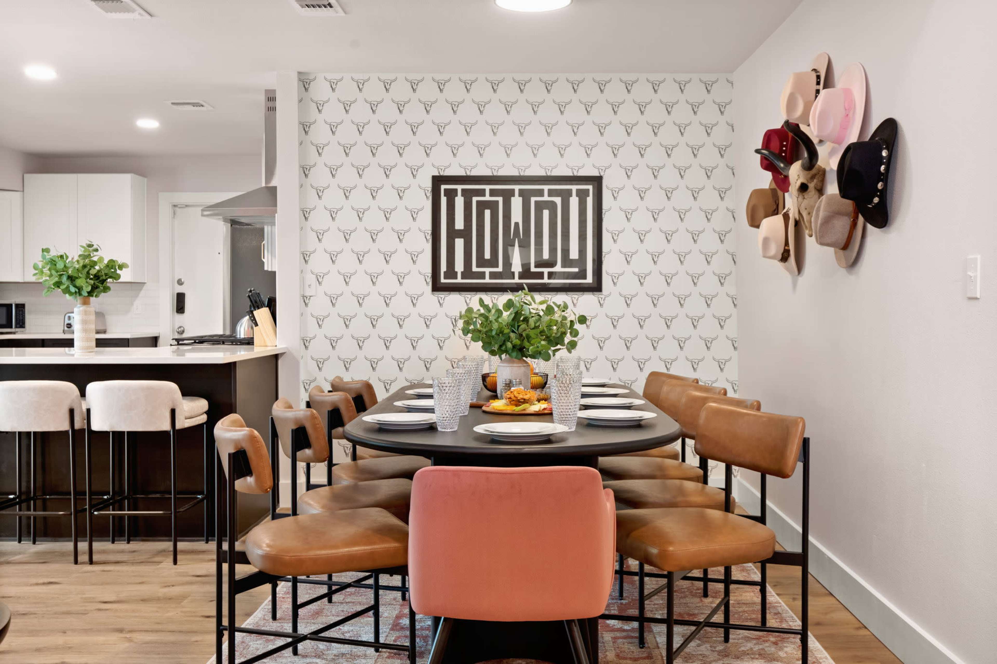 A dining area features a round black table surrounded by eight brown and pink chairs, with a decorative wall showcasing a "HOWDY" sign and a collection of hats.