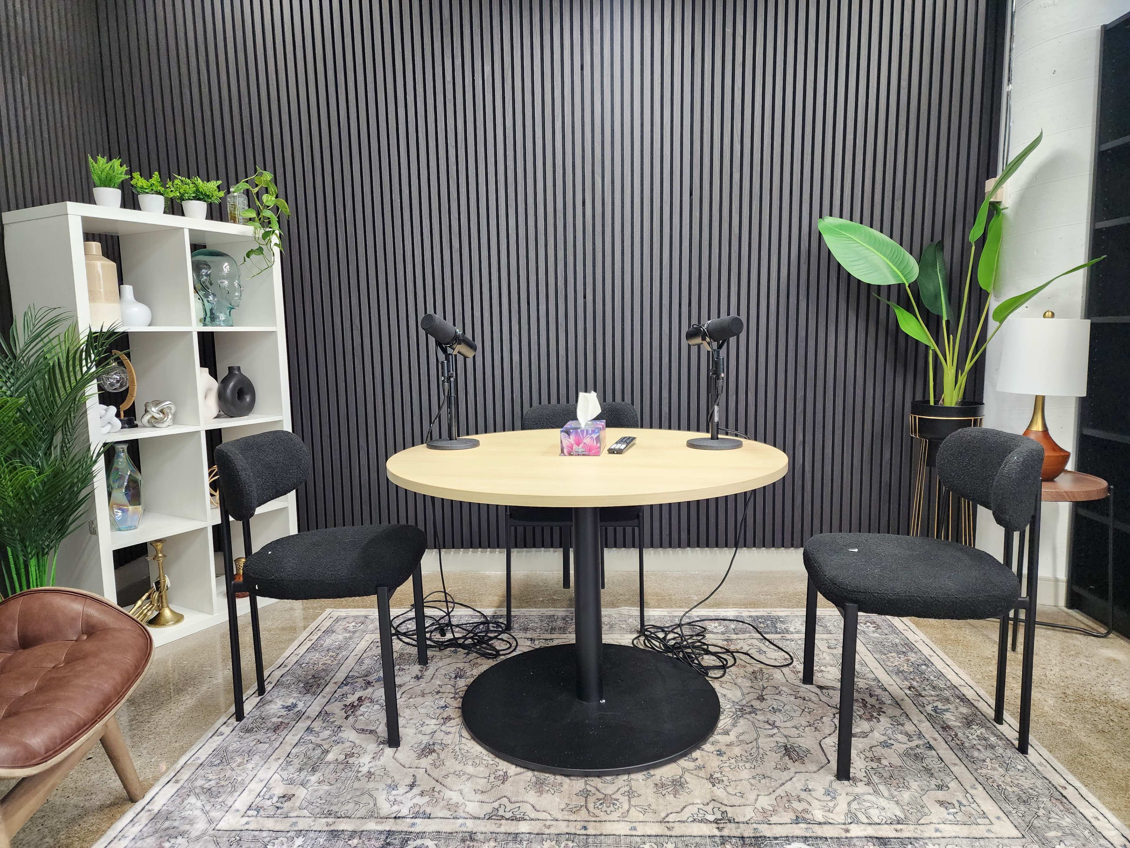 The image shows a podcast recording setup featuring a round table with three chairs, two microphones, and a background of textured black panels and green plants.