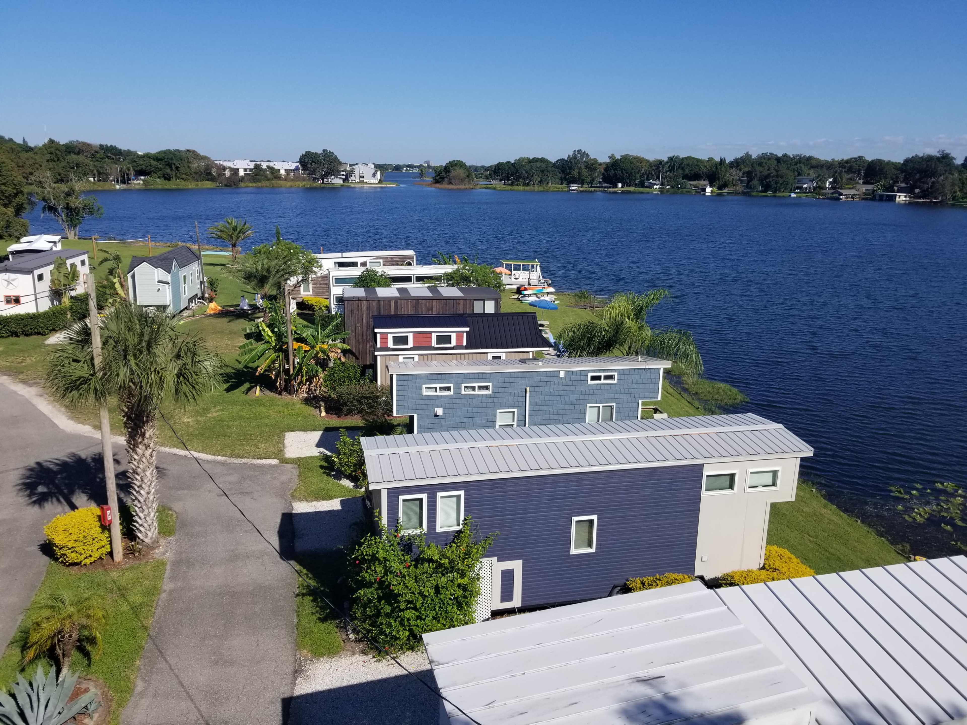 A row of colorful tiny homes along a waterfront with a clear blue sky and lush greenery.