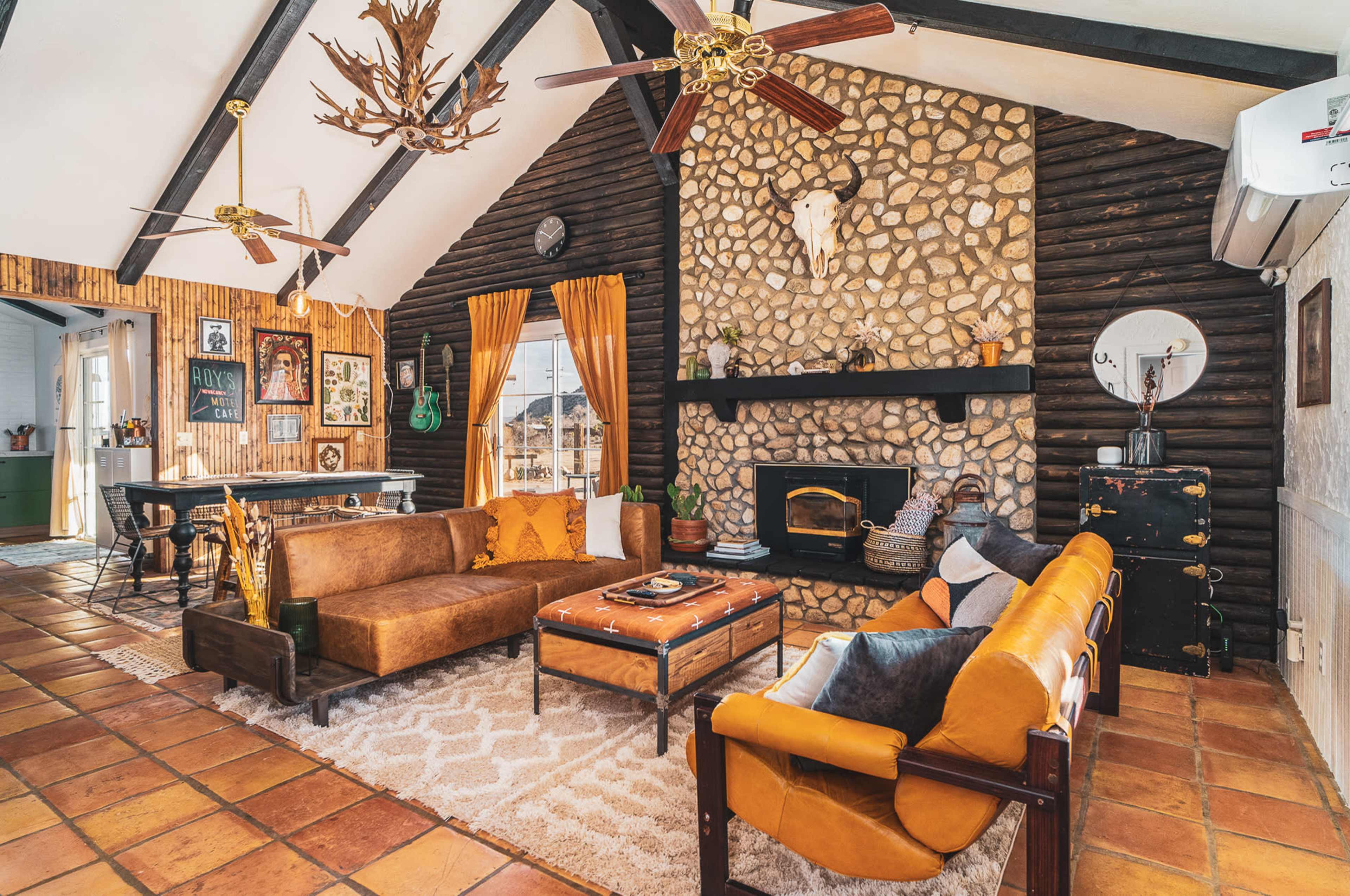 The living room features a stone fireplace, wooden beams, and a mix of brown and orange sofas arranged around a coffee table on a patterned rug.