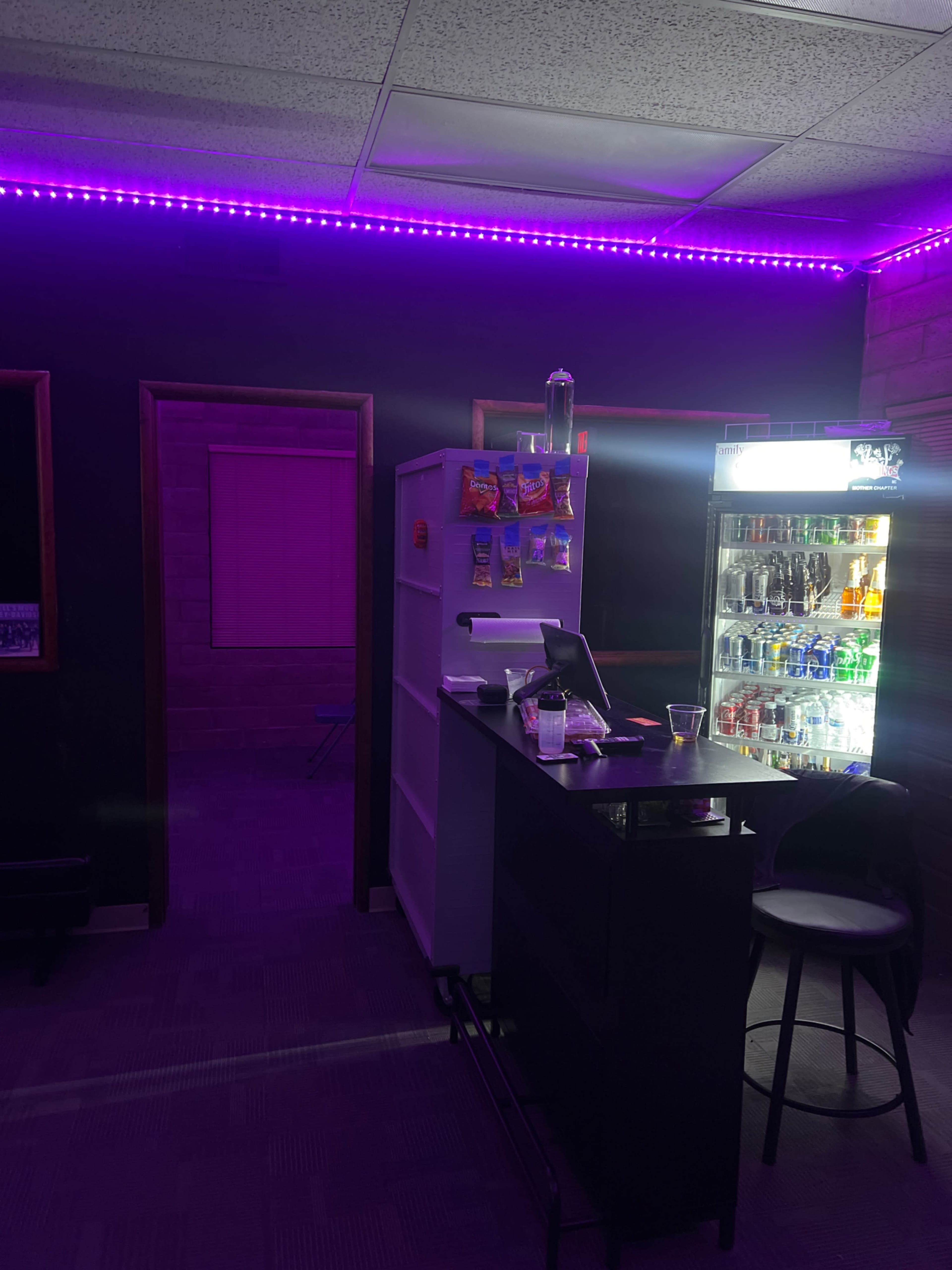 The image shows a dimly lit room with purple LED lighting, featuring a bar area with a cash register and a refrigerator stocked with beverages.