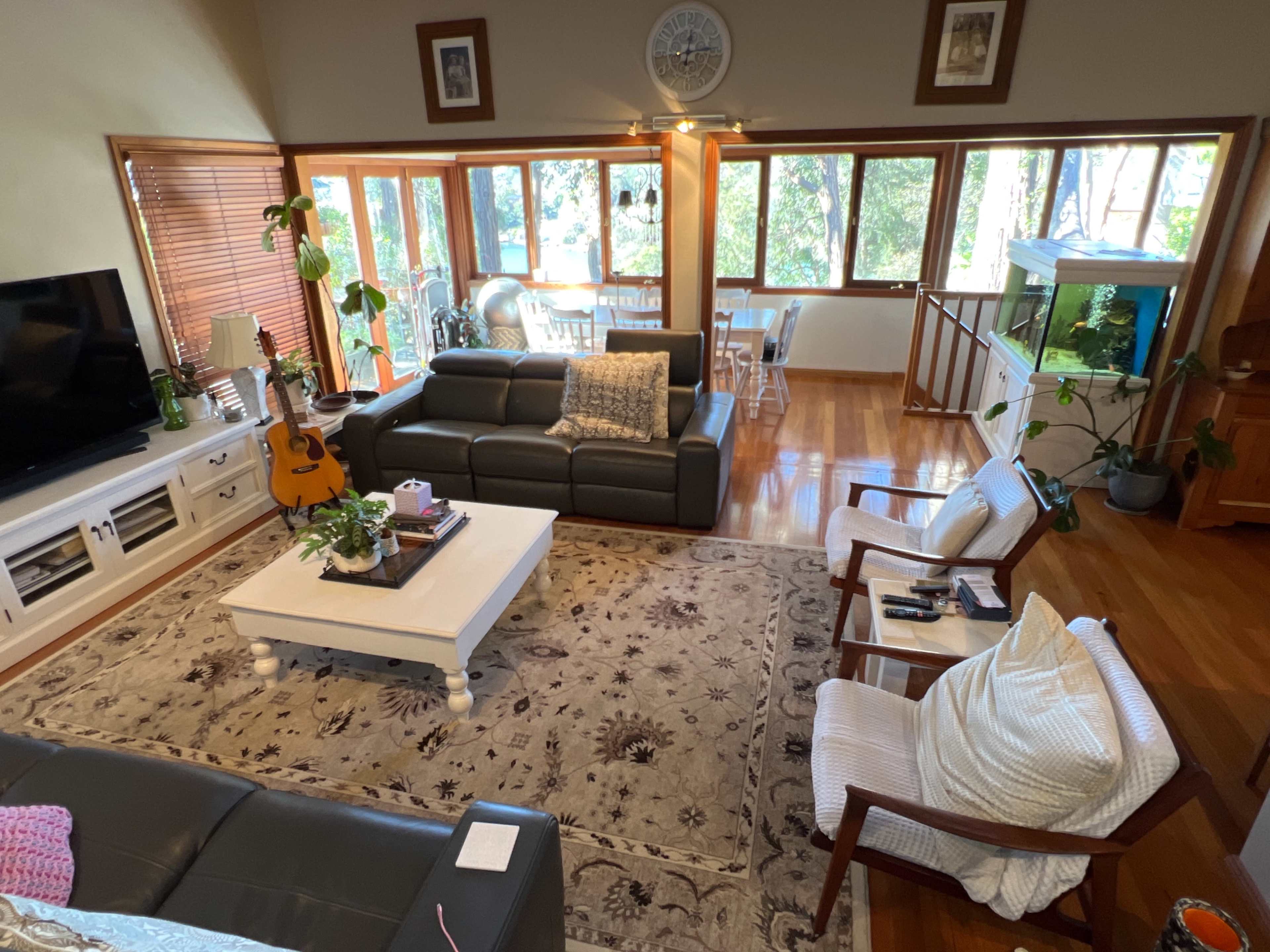 A spacious living area with a dark leather sofa, a white coffee table, an acoustic guitar, and large windows overlooking a natural setting.