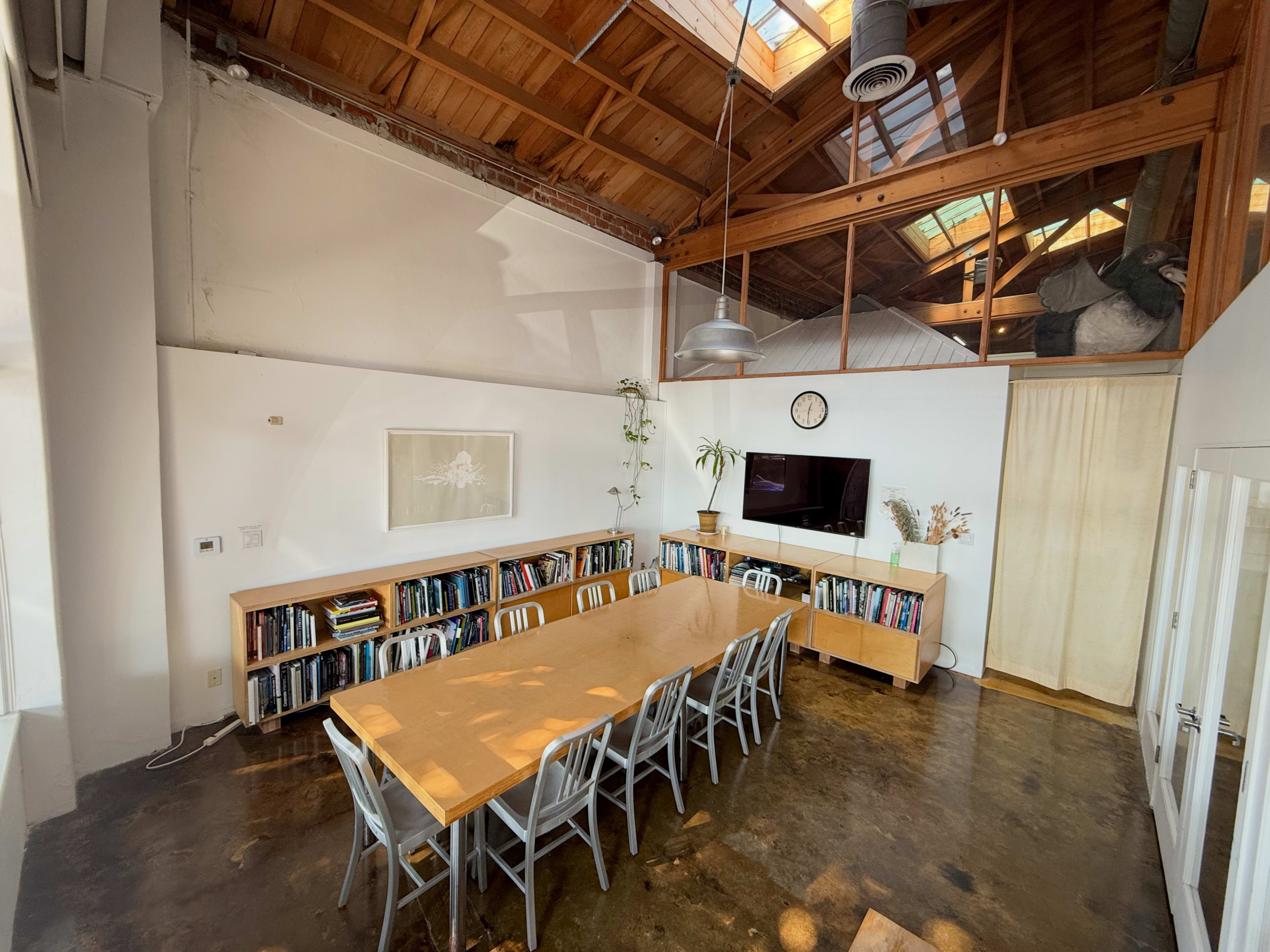 The image shows a spacious meeting room with a long wooden table, surrounded by chairs, and shelves filled with books along one wall.