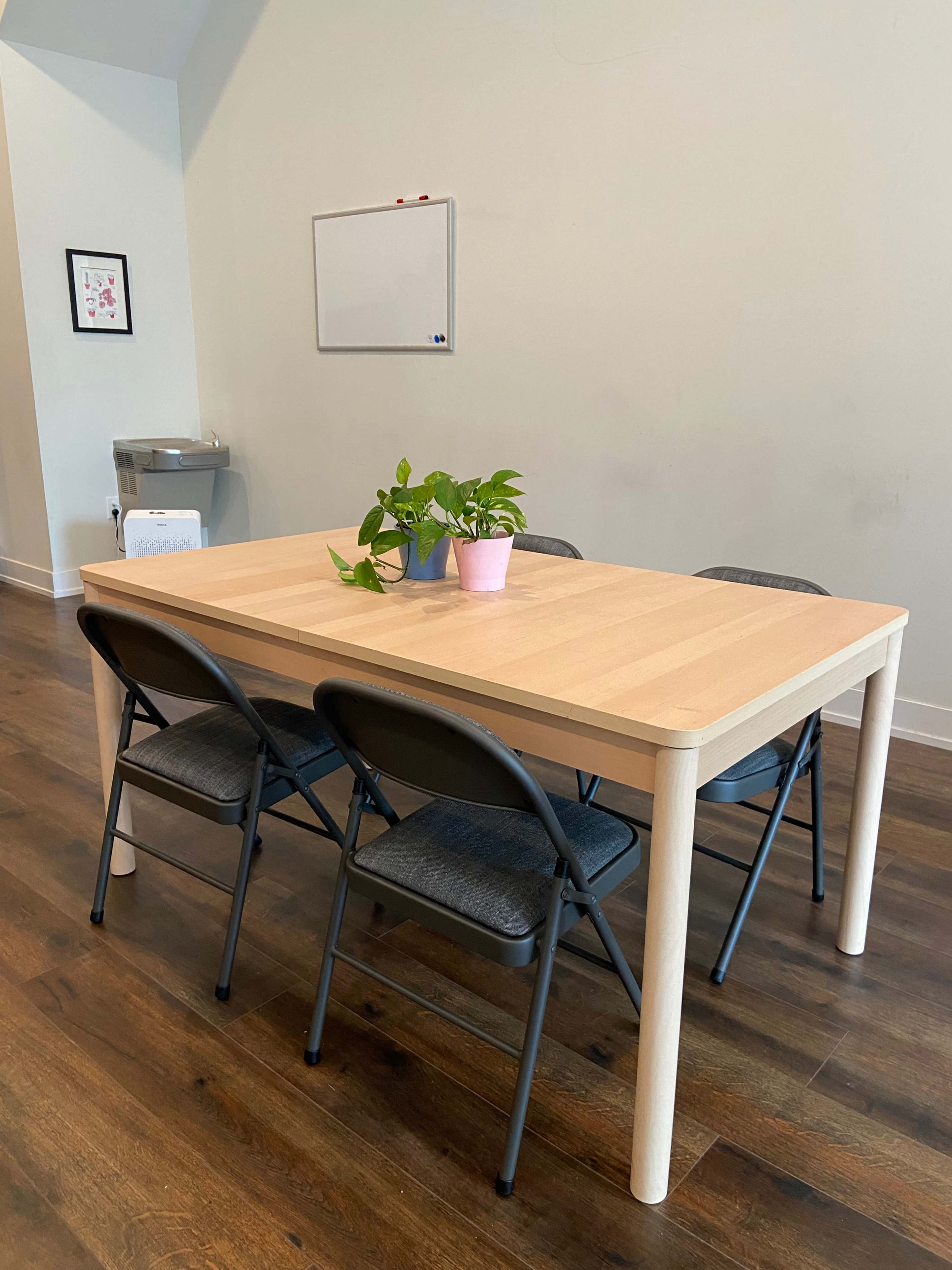 A wooden table with four black folding chairs is set in a room with a small plant in a pink pot at its center.