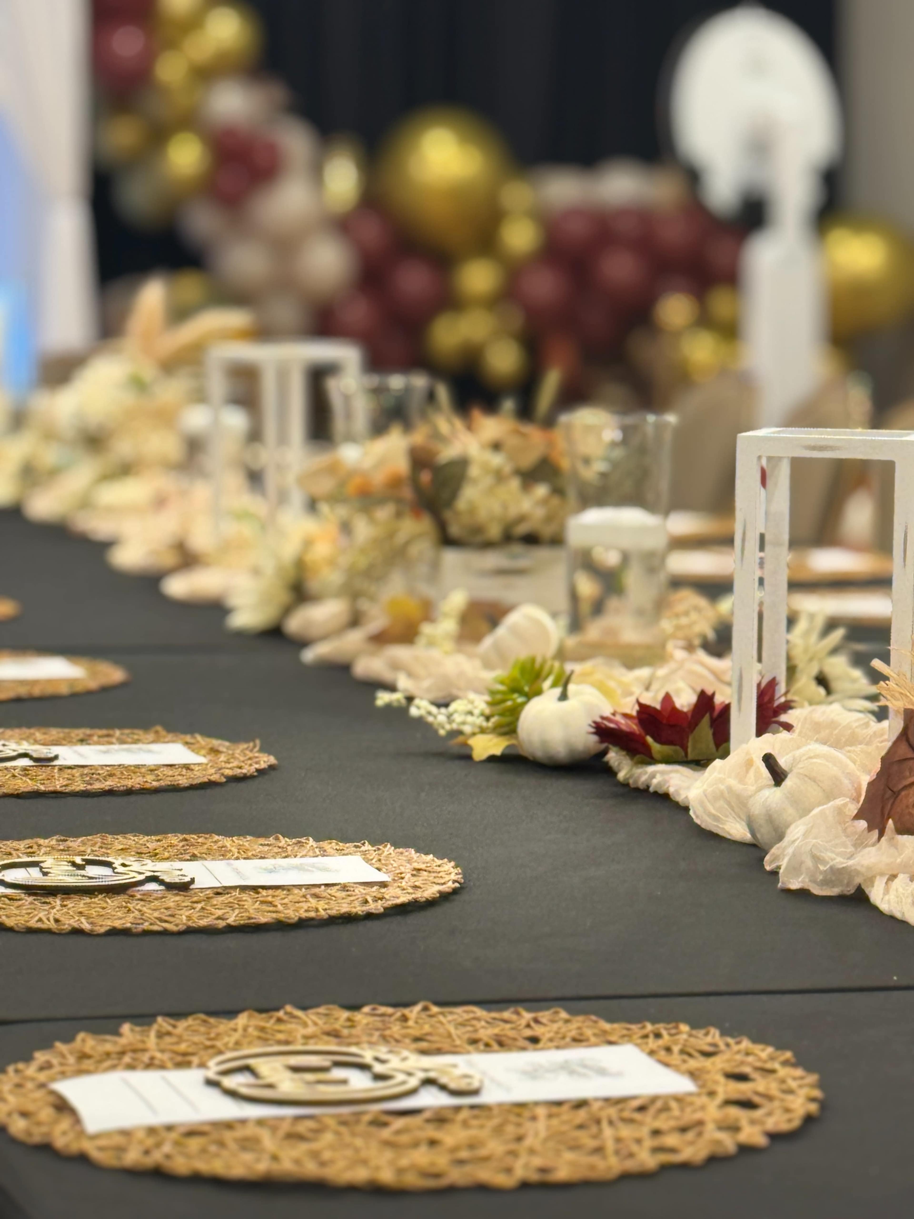 A long banquet table is decorated with woven placemats, floral arrangements, and candles, set against a backdrop of balloons in gold and burgundy.