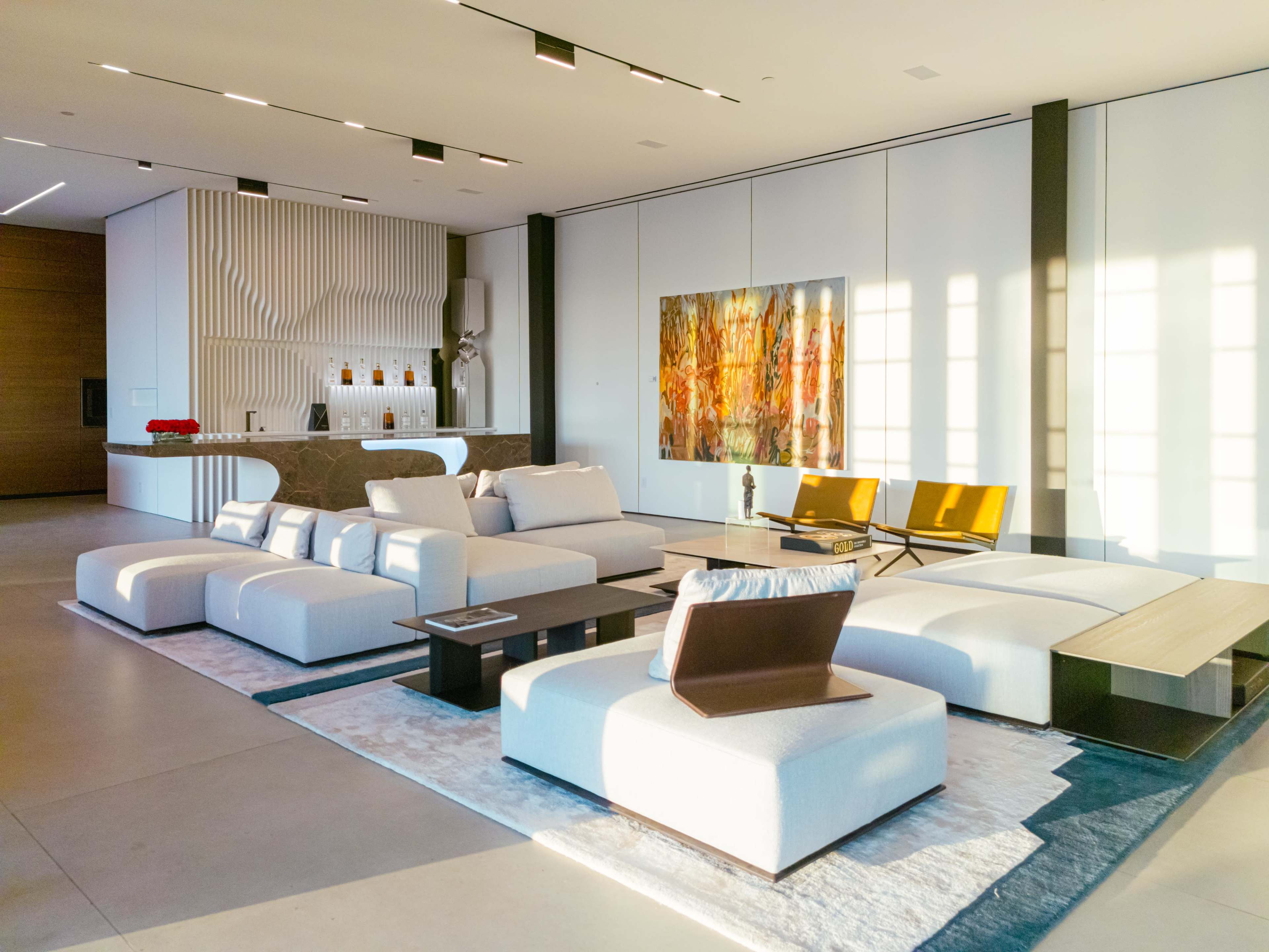 A modern, minimalist living room with white sofas, a wooden coffee table, an abstract painting on the wall, and a kitchen area in the background.