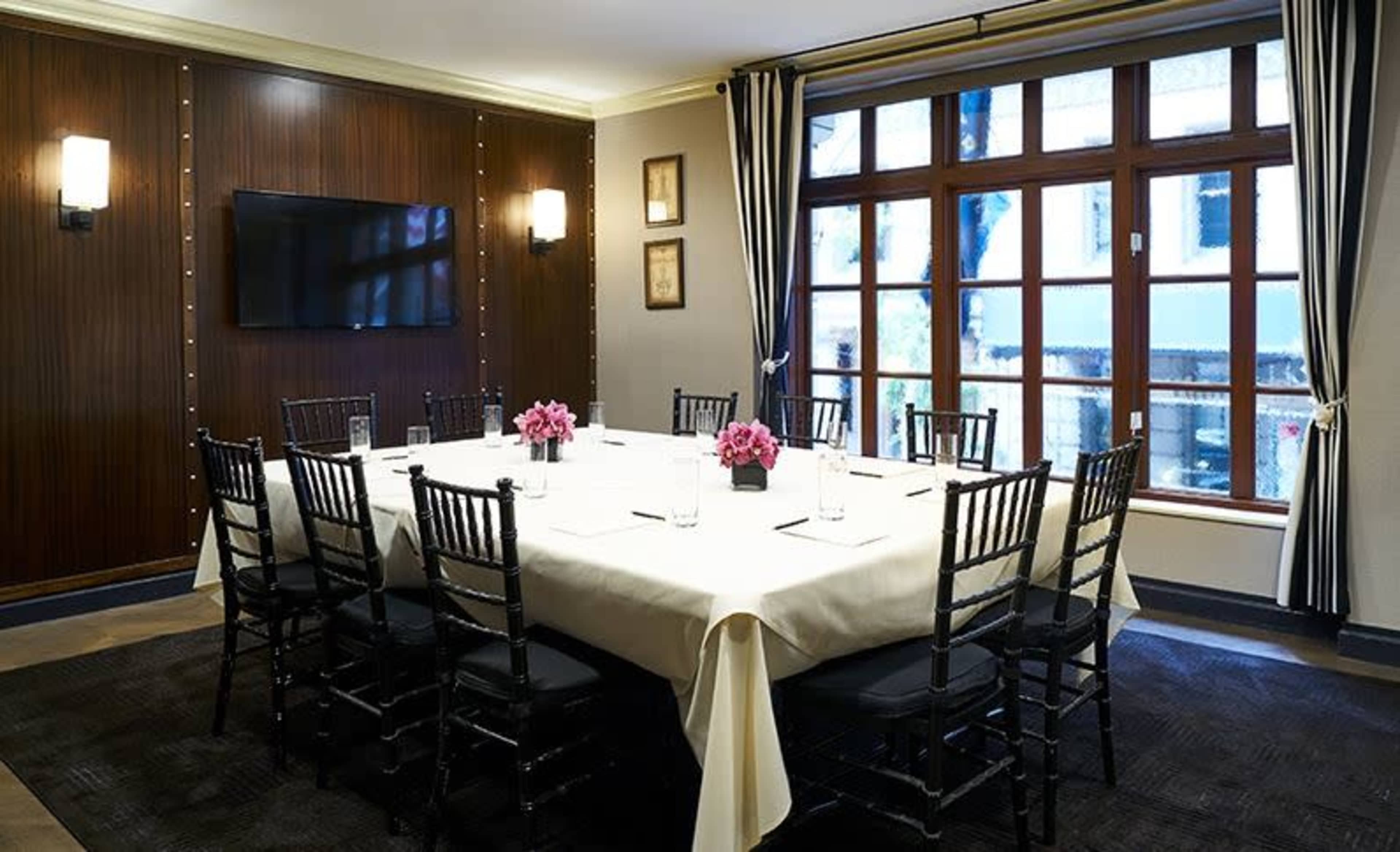A conference room features a large, rectangular table surrounded by black chairs, with a floral centerpiece and a large window providing natural light.