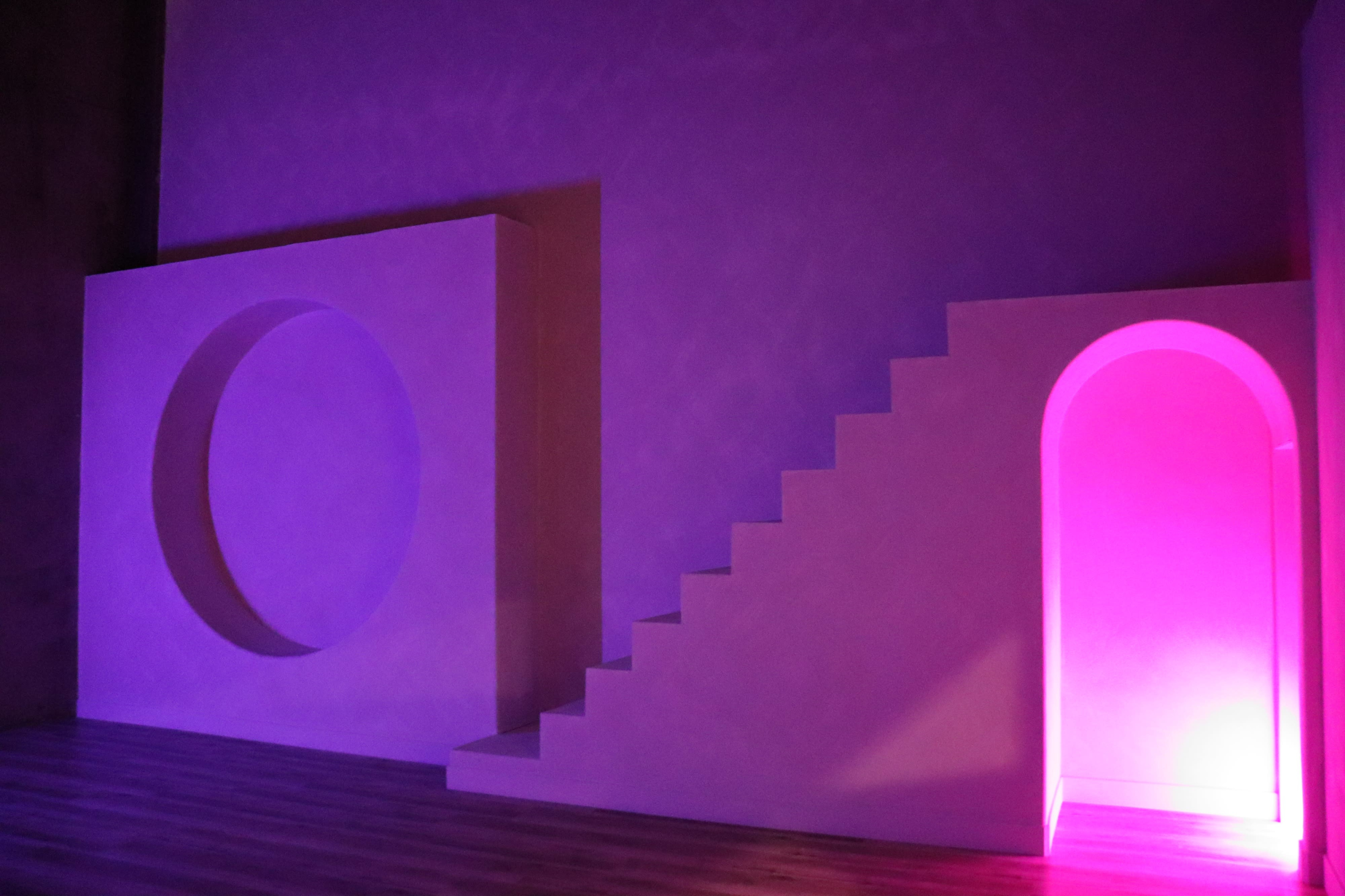 The image depicts a minimalist interior featuring a curved wall segment, a staircase, and an arched doorway, illuminated with a purple light.