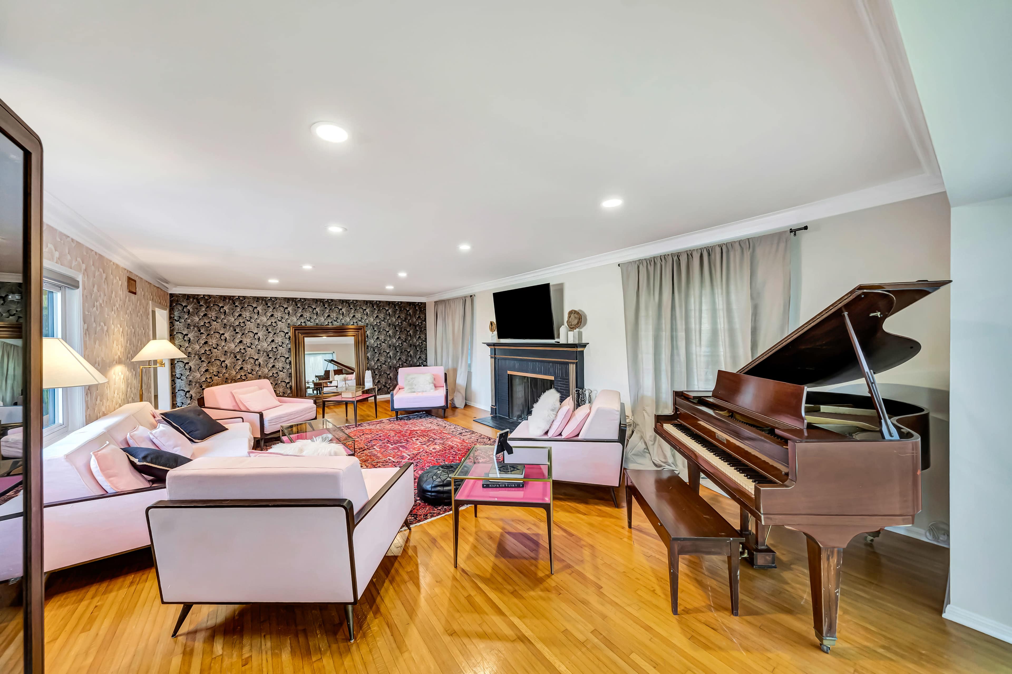 A spacious living room features a grand piano, pink upholstered furniture, a large mirror, and a TV mounted above a fireplace.