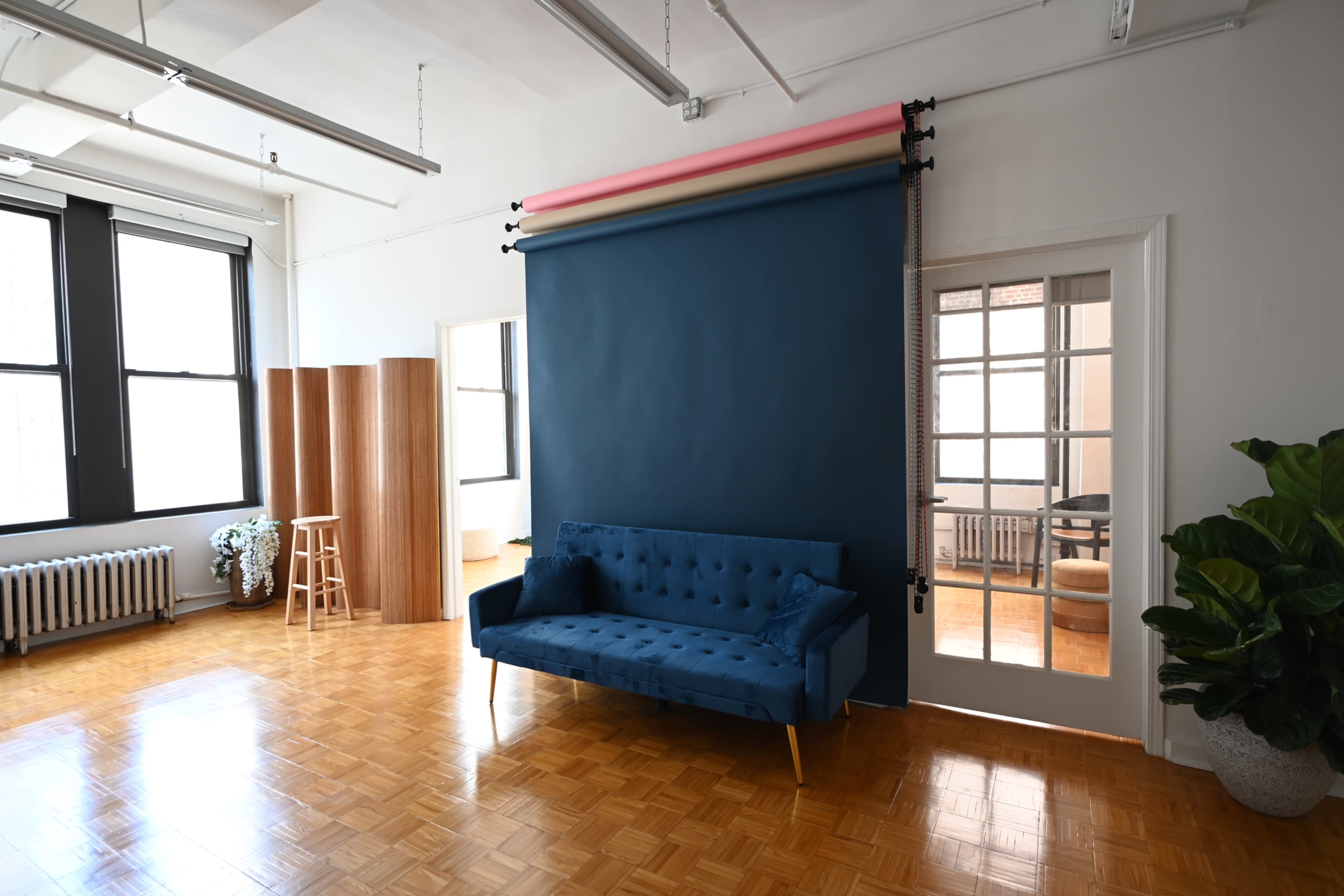 The image shows a spacious room with a blue backdrop, a blue upholstered couch, wooden furniture, and large windows allowing natural light to enter.
