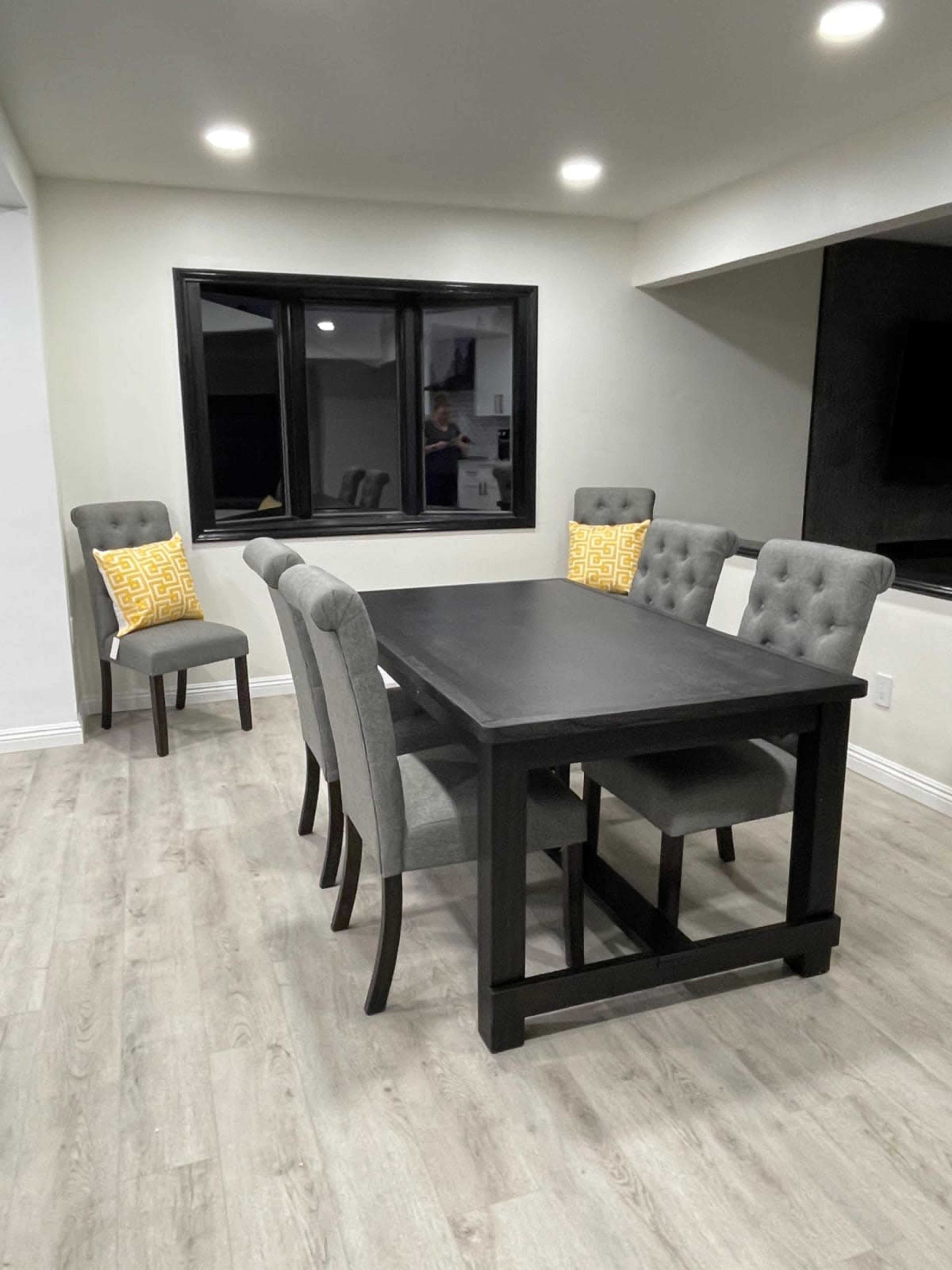 A rectangular dining table is surrounded by six chairs, with two adorned with yellow patterned cushions, set in a well-lit room with a large window.