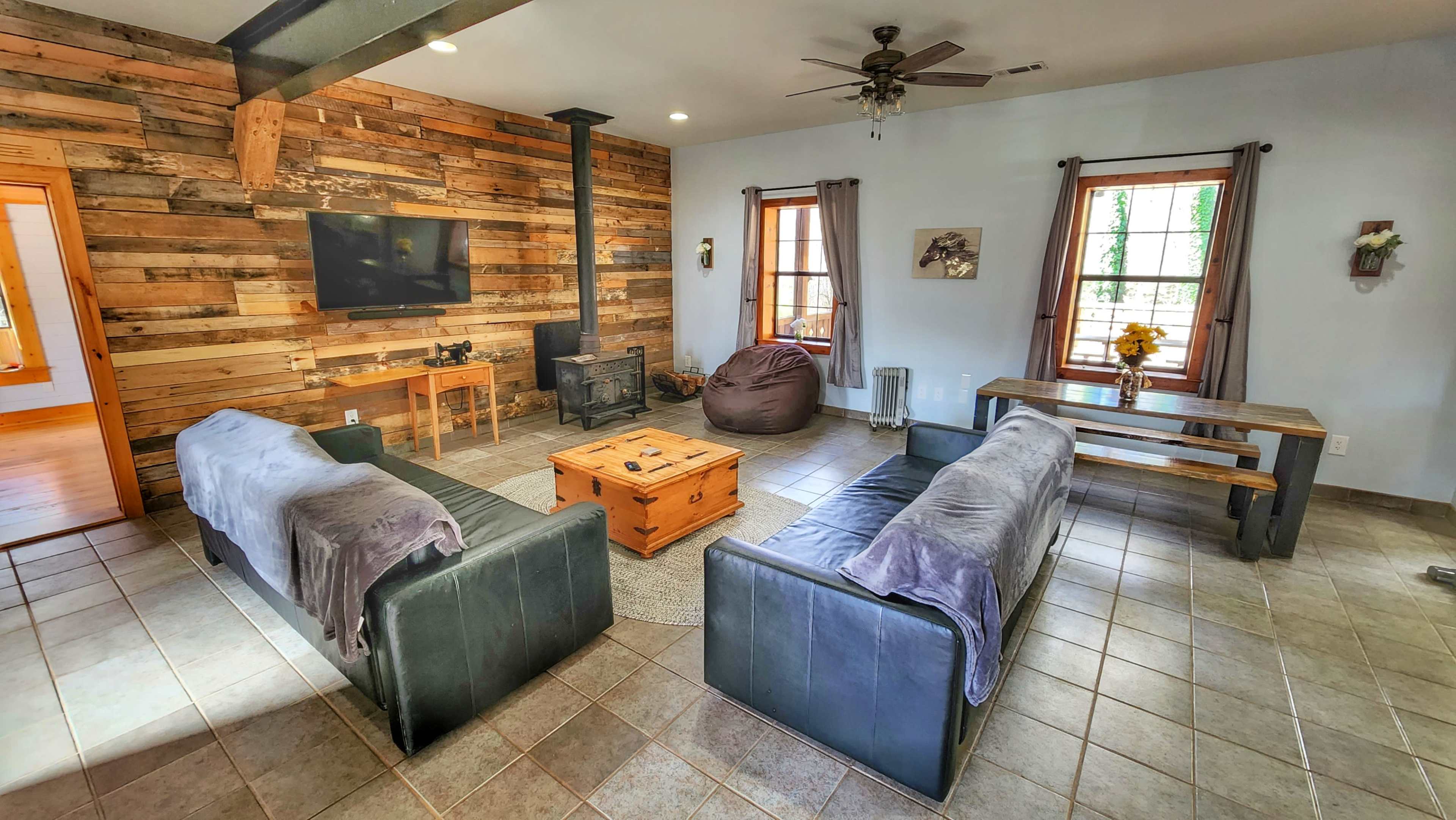 The living room features a wooden accent wall, two sofas facing a central coffee table, and a stove, with natural light streaming through the windows.