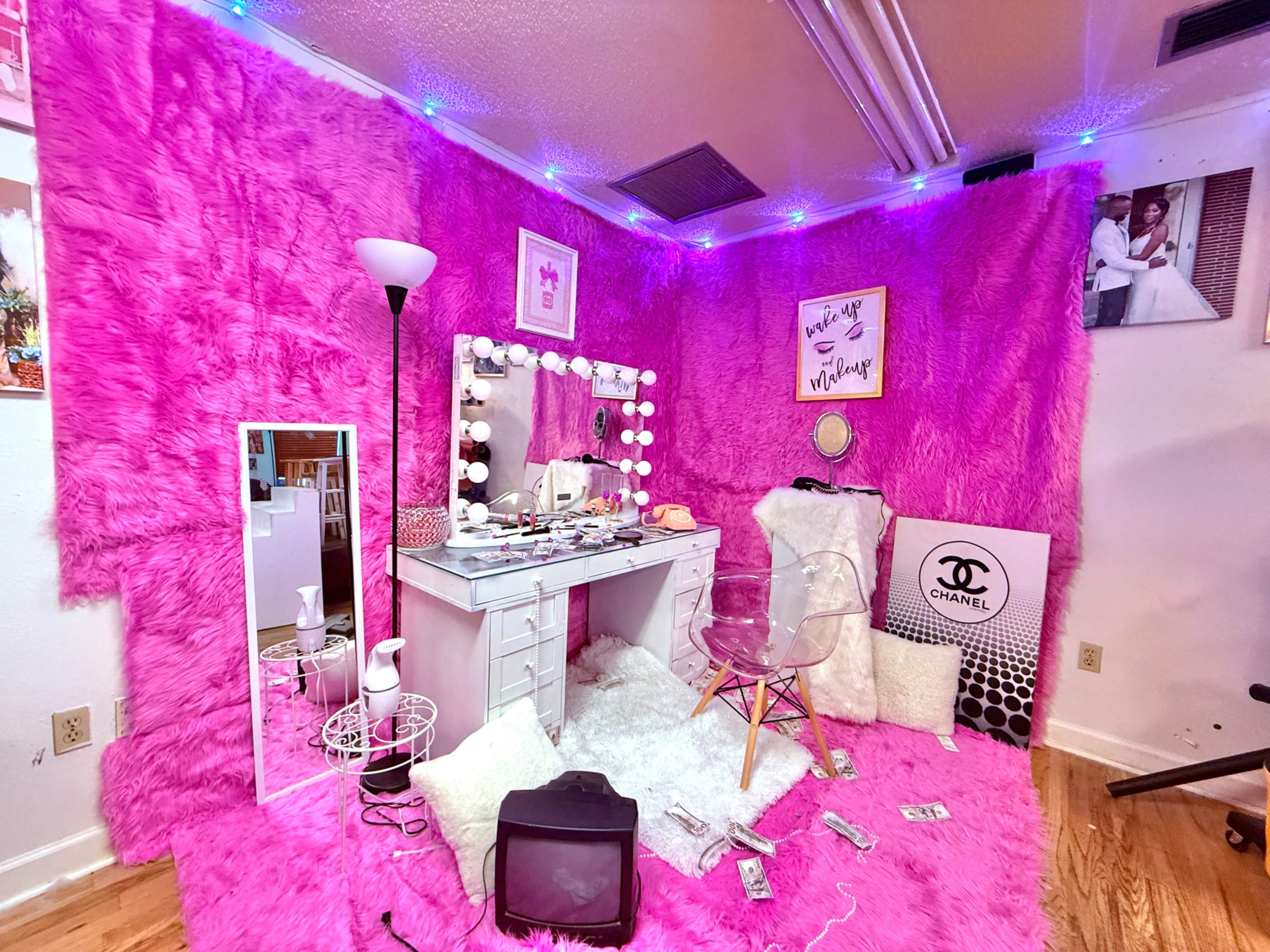 The image shows a brightly colored pink room with fuzzy pink walls, a vanity table with a mirror and lights, and various items scattered around, including a vintage television and a white chair.