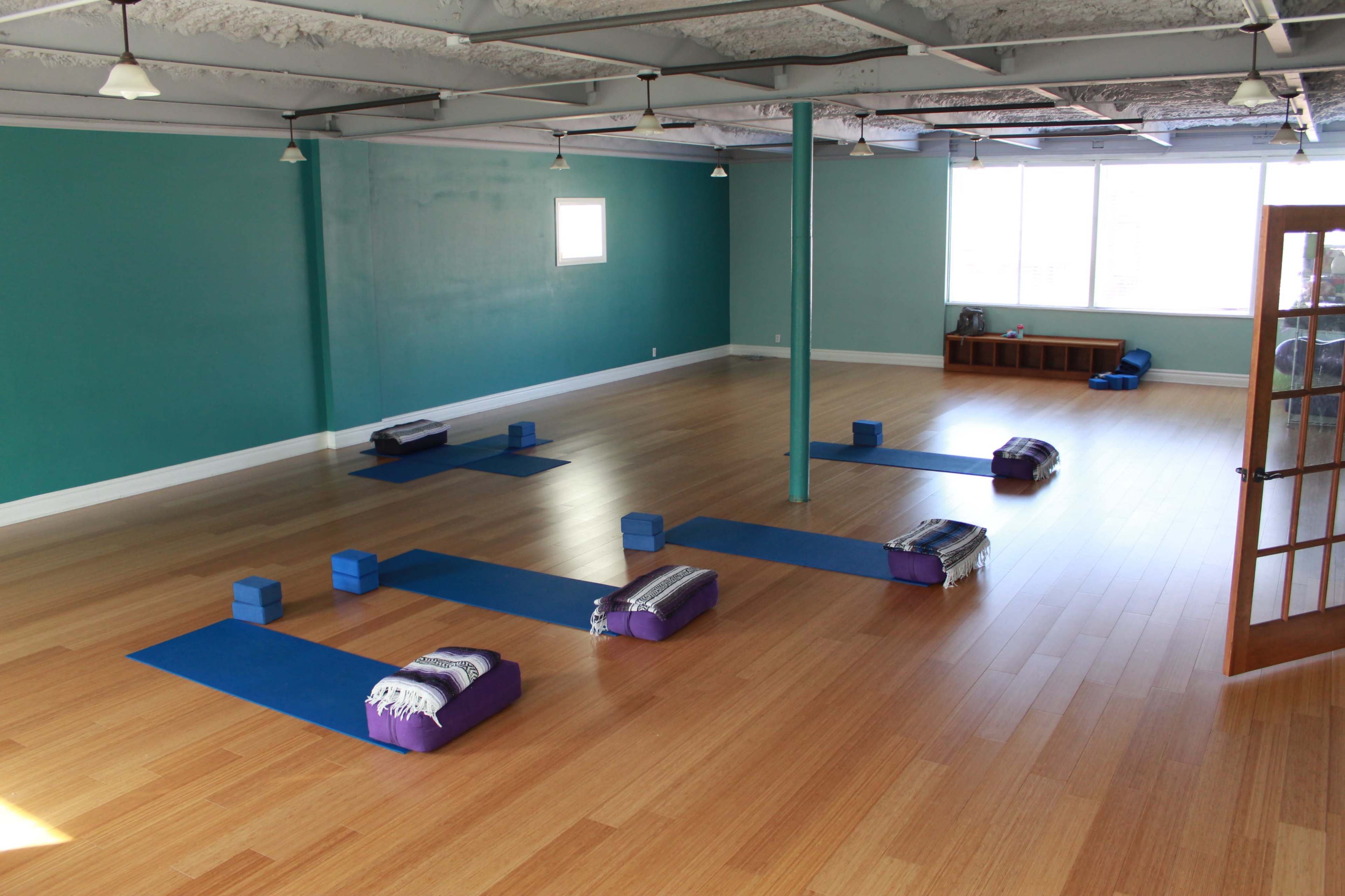 The image shows a yoga studio with polished wooden floors, blue yoga mats arranged in a specific pattern, and several purple bolsters and blocks placed on the mats.