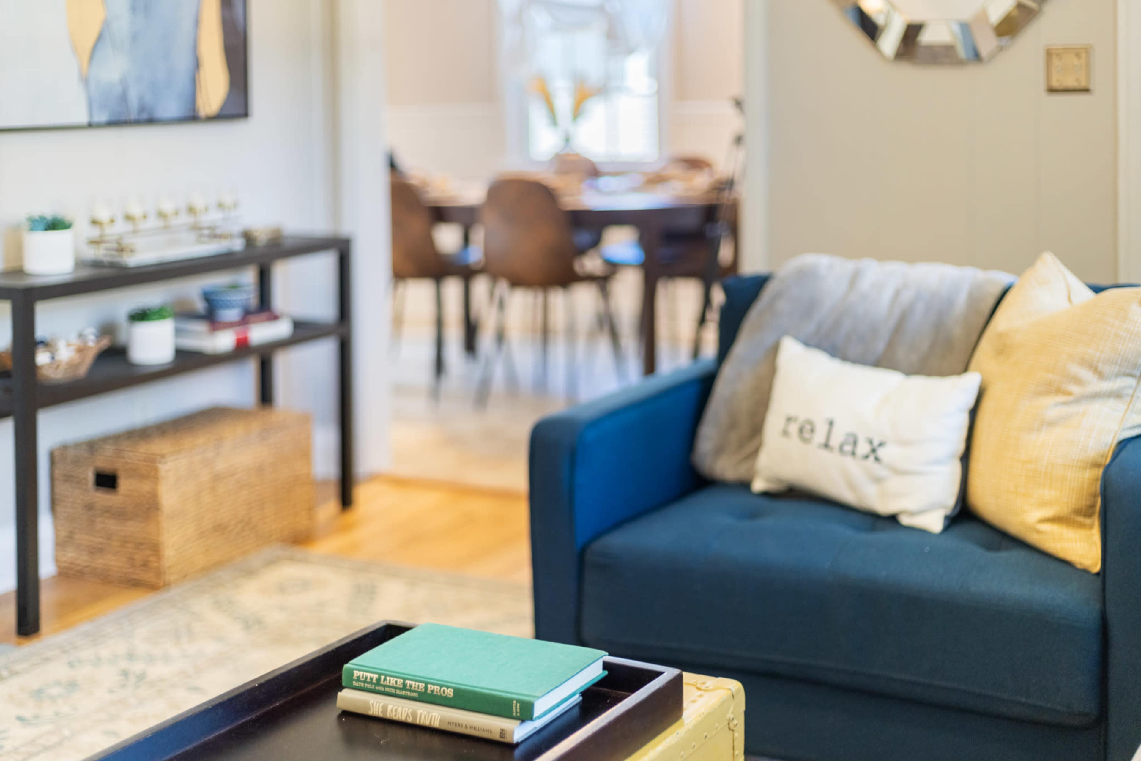 A blue couch with a "relax" pillow sits in a living area, with a coffee table holding books, and a dining room visible in the background.