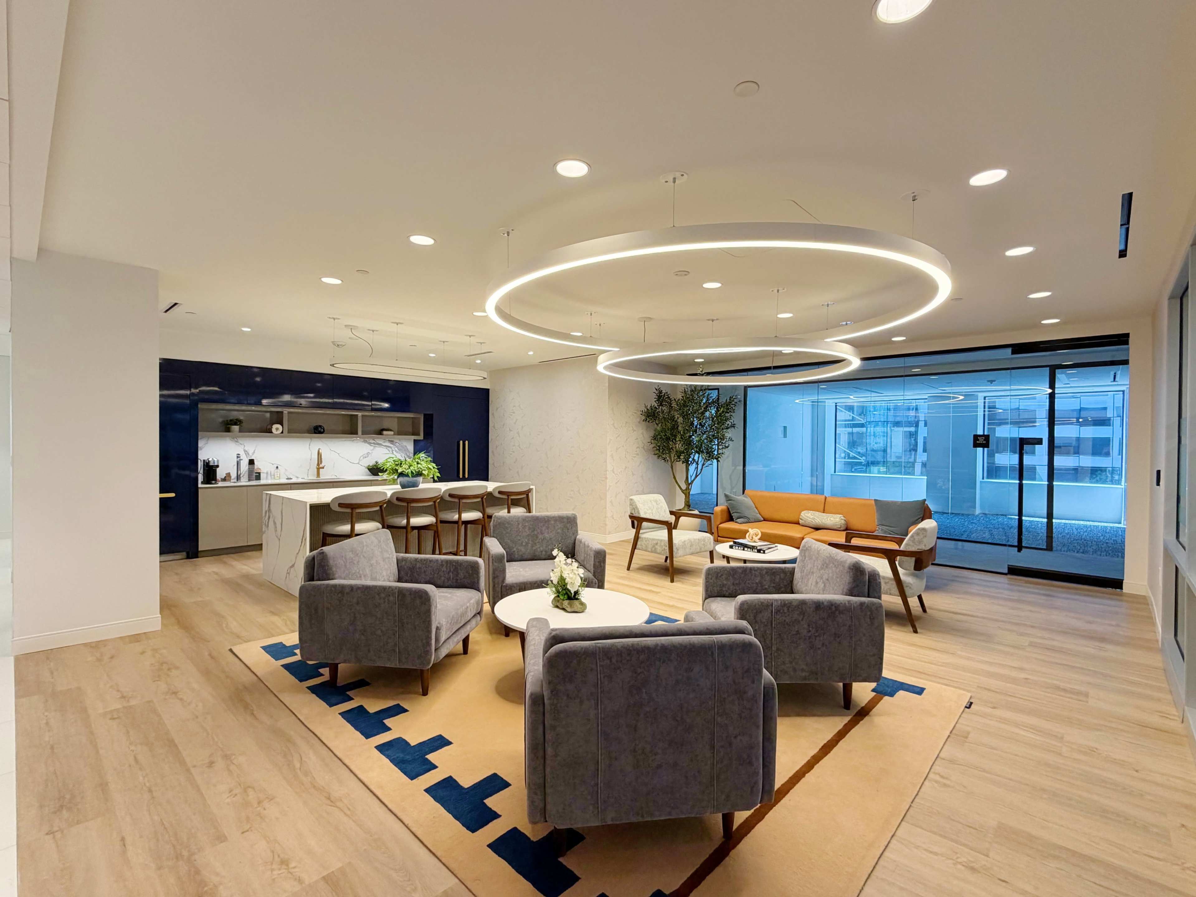 The image shows a modern office lounge area featuring a seating arrangement with gray armchairs and an orange sofa, a kitchen space in the background, and large windows.