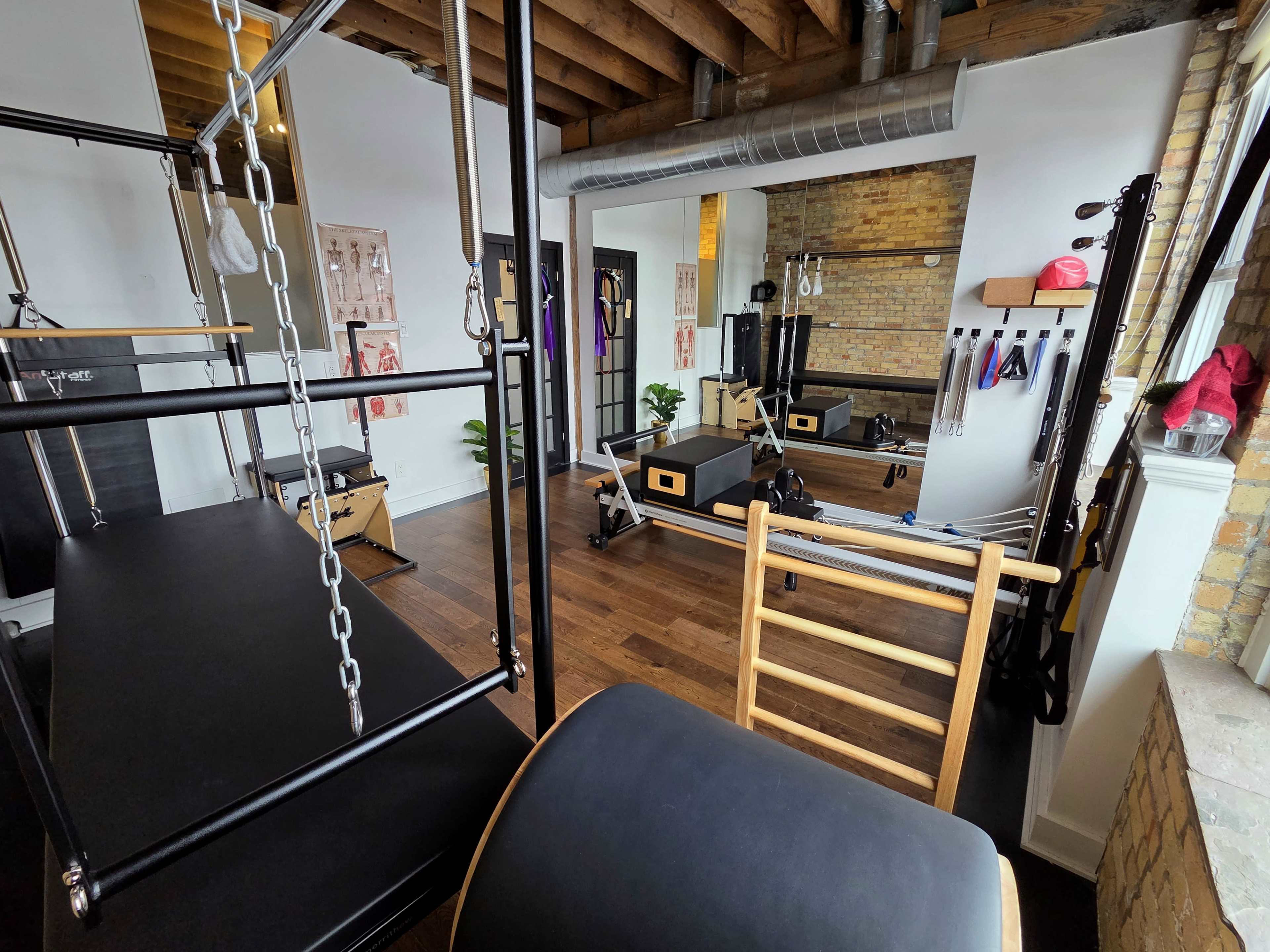 The image shows a well-lit Pilates studio featuring various equipment, including reformers, a ladder barrel, and wall-mounted accessories, within a modern interior space.