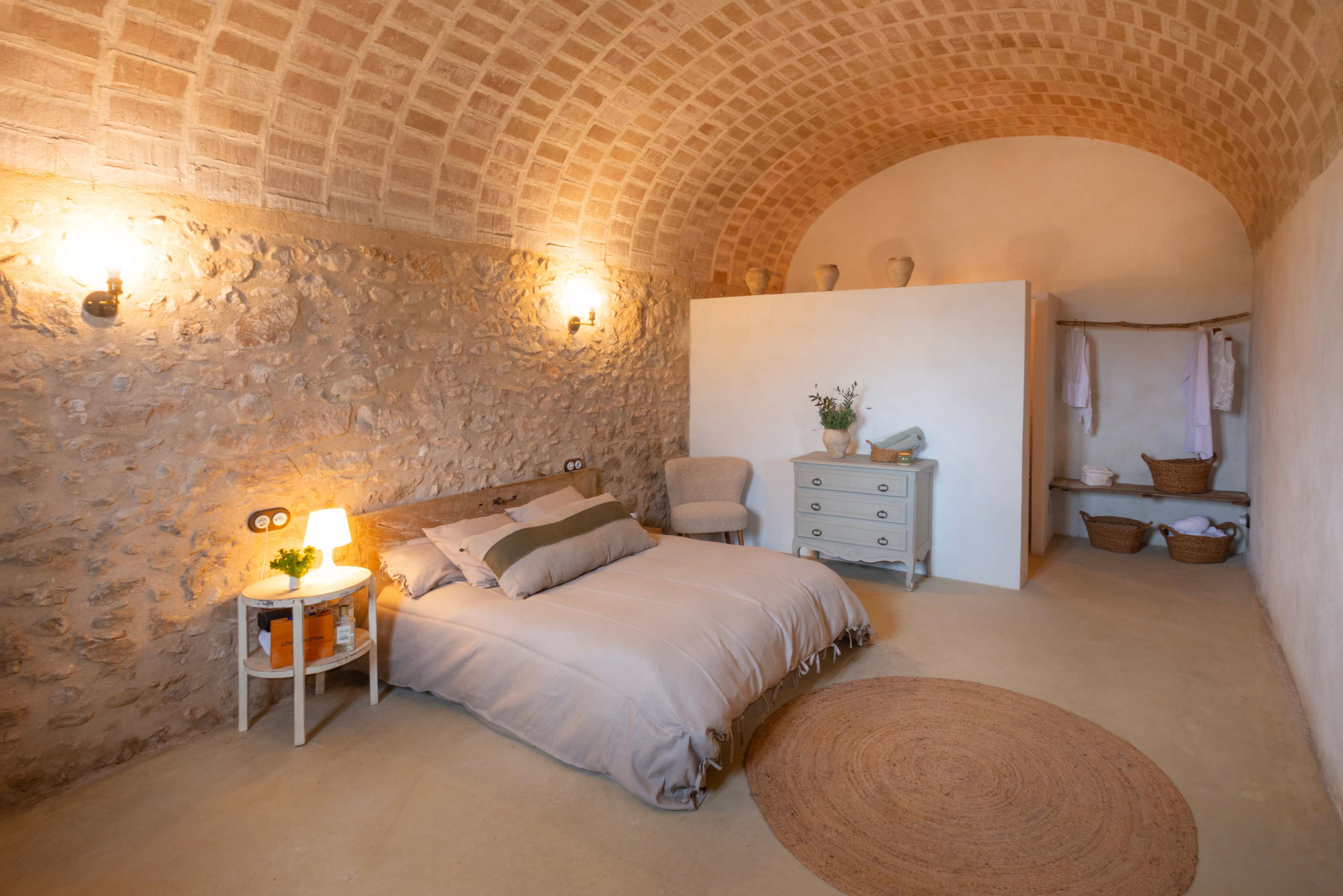 The image shows a minimalist bedroom with a stone wall, an arched ceiling, a bed, a small nightstand with a lamp, and a dresser, complemented by a cozy rug on the floor.