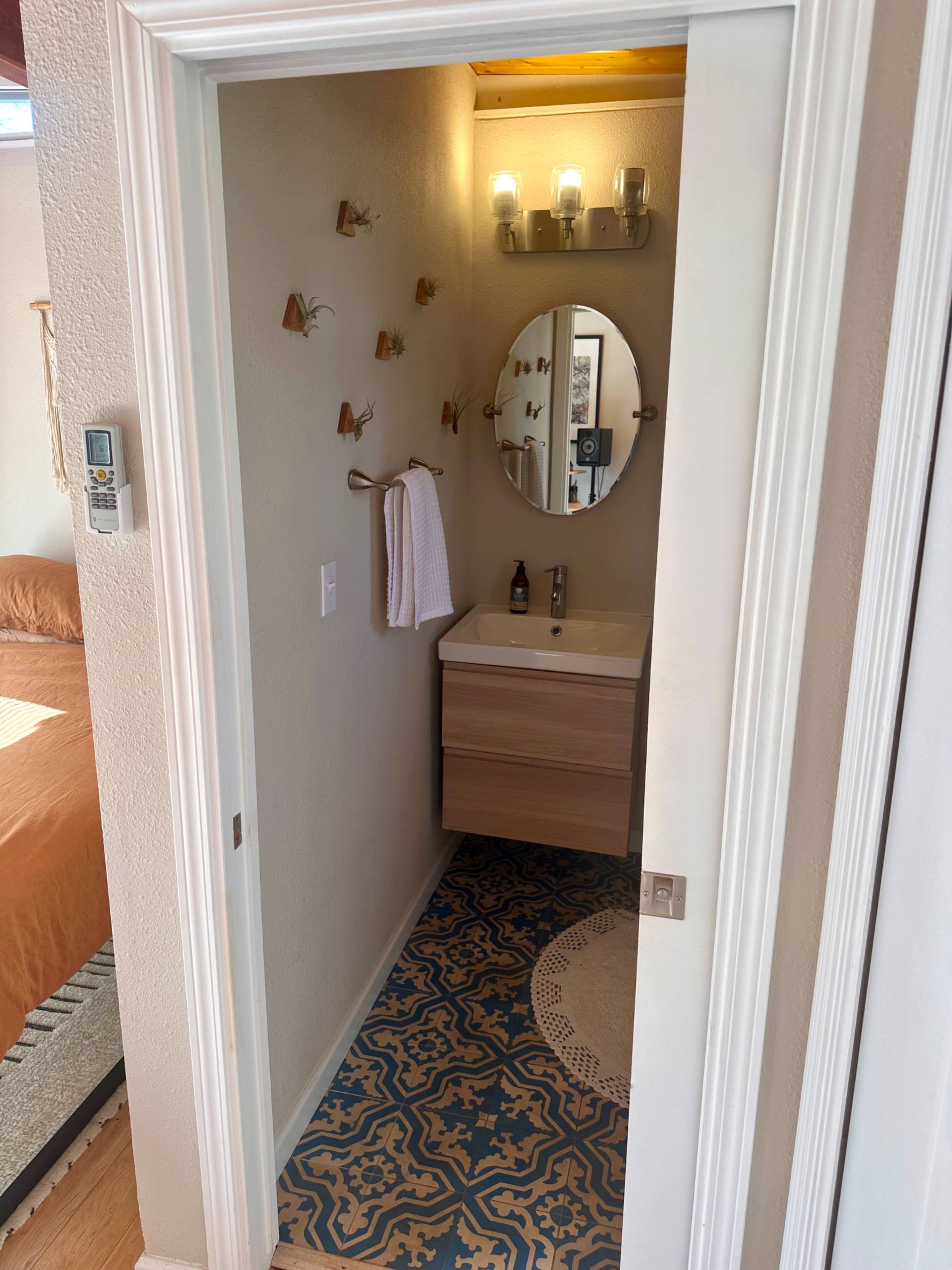 The image shows a bathroom entrance featuring a wooden vanity with a round mirror, a wall-mounted towel rack, and decorative plants on the wall, with patterned flooring visible.