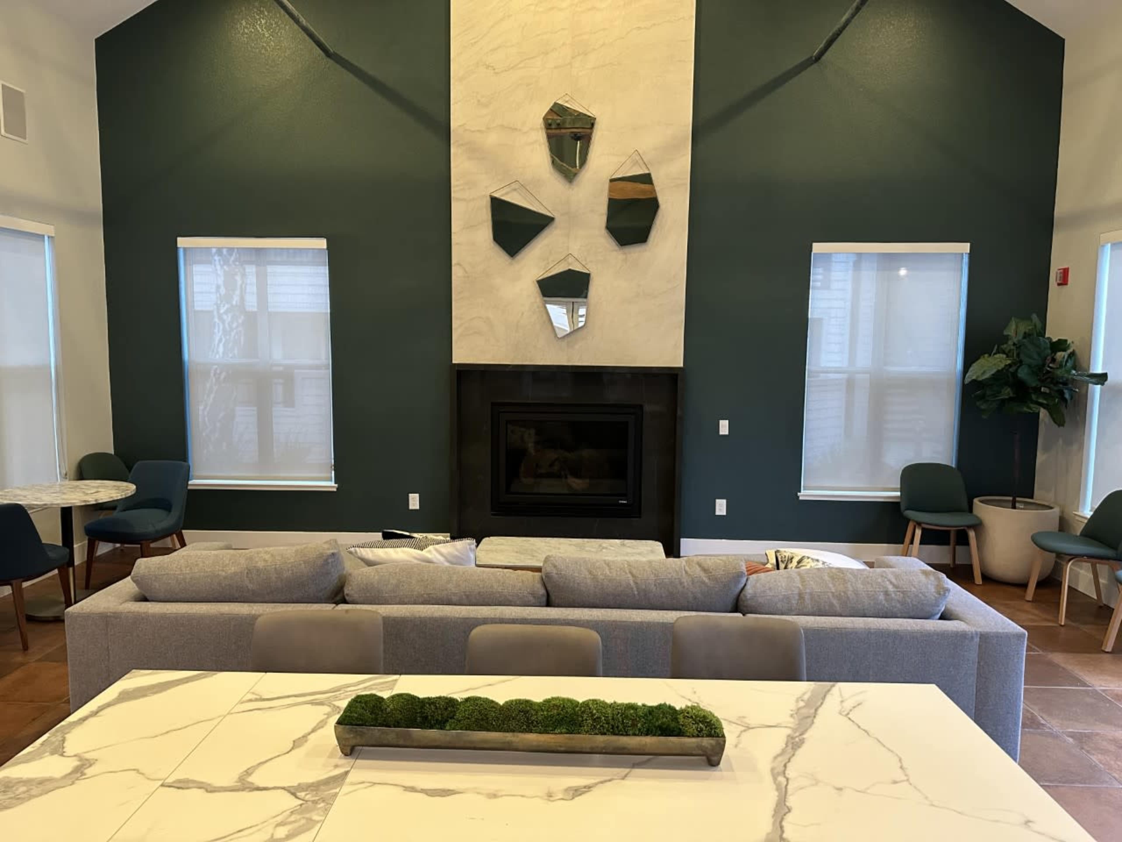 A modern living area with a gray sofa facing a minimalist fireplace, framed by geometric mirrors on the stone wall, and surrounded by green accent chairs and tables.