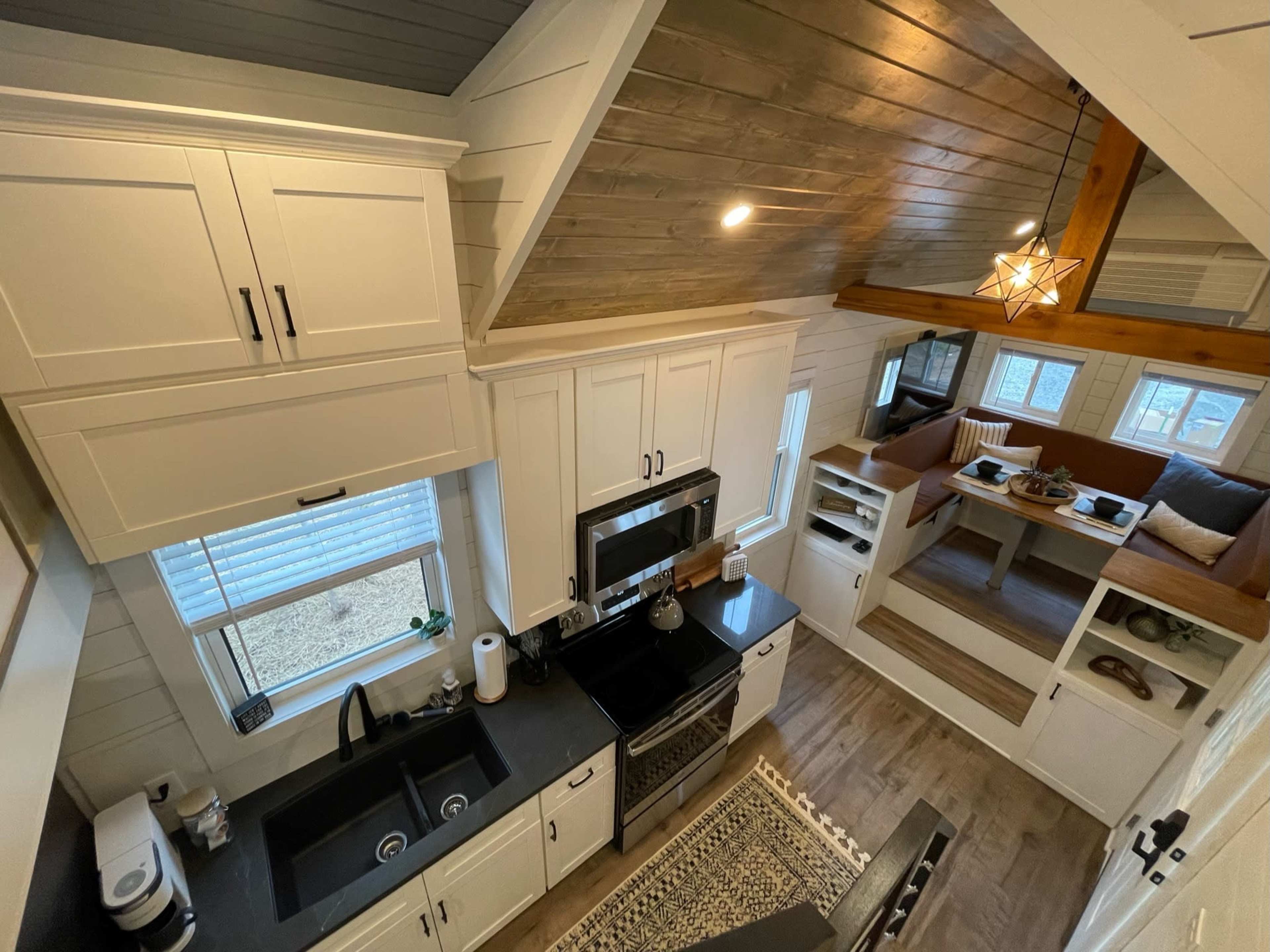 A small, modern kitchen with white cabinetry, black countertops, and a cozy seating area featuring built-in shelves and large windows.