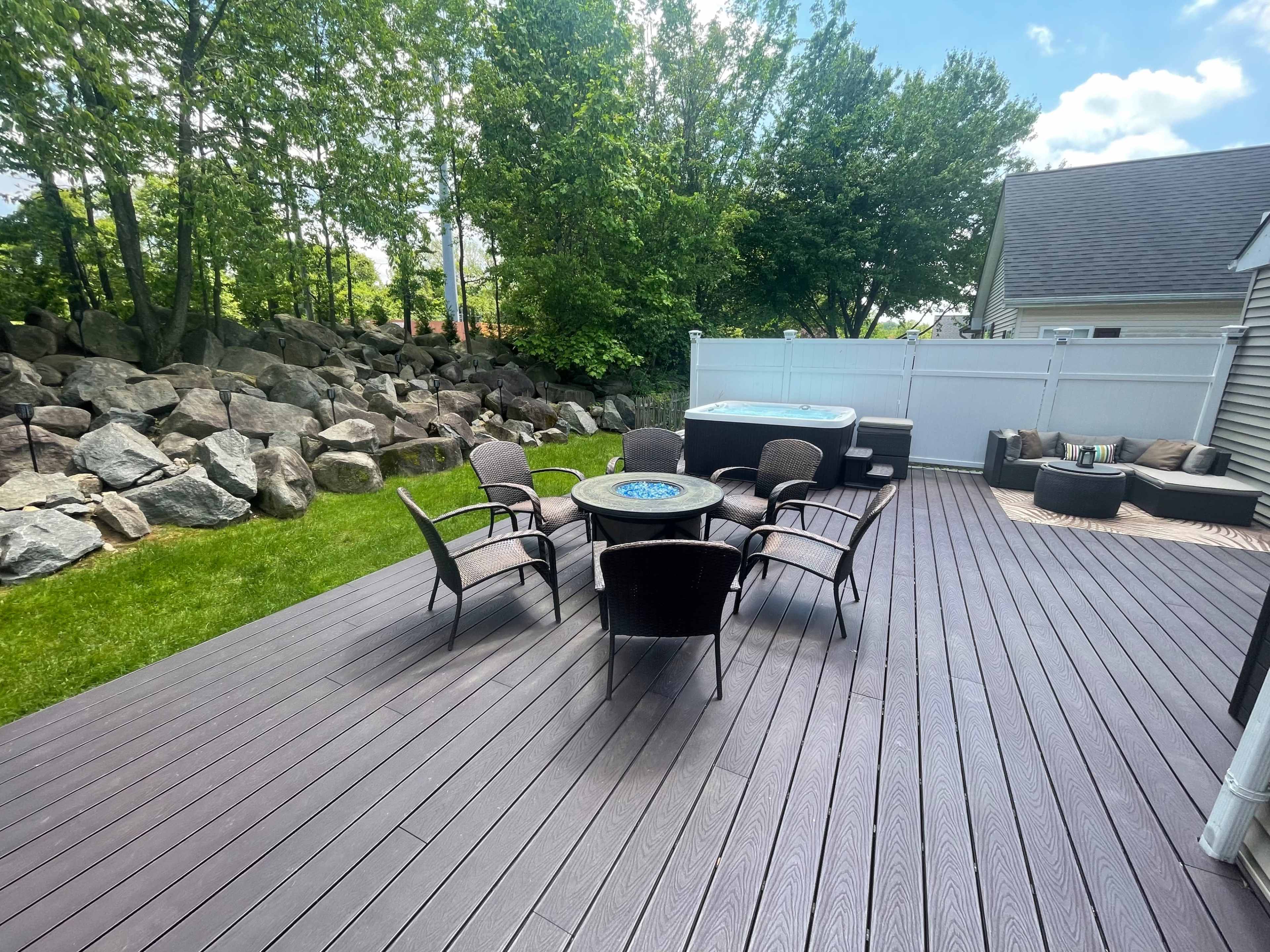 A wooden deck with multiple chairs arranged around a circular table, surrounded by trees and a rocky landscape in the background.