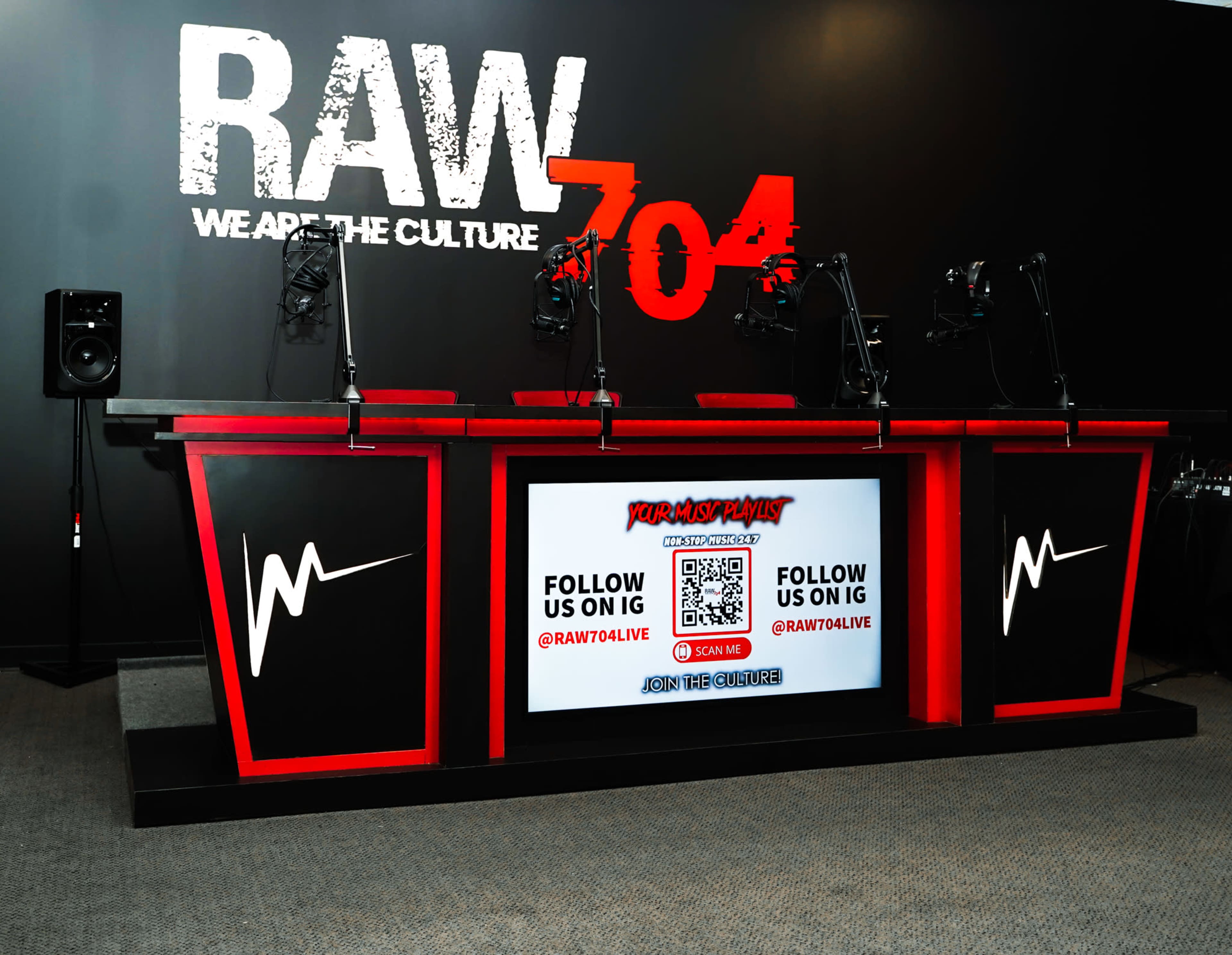 A modern studio setup with a red and black broadcast table featuring multiple microphones and a large screen displaying social media information against a black wall with bold text.
