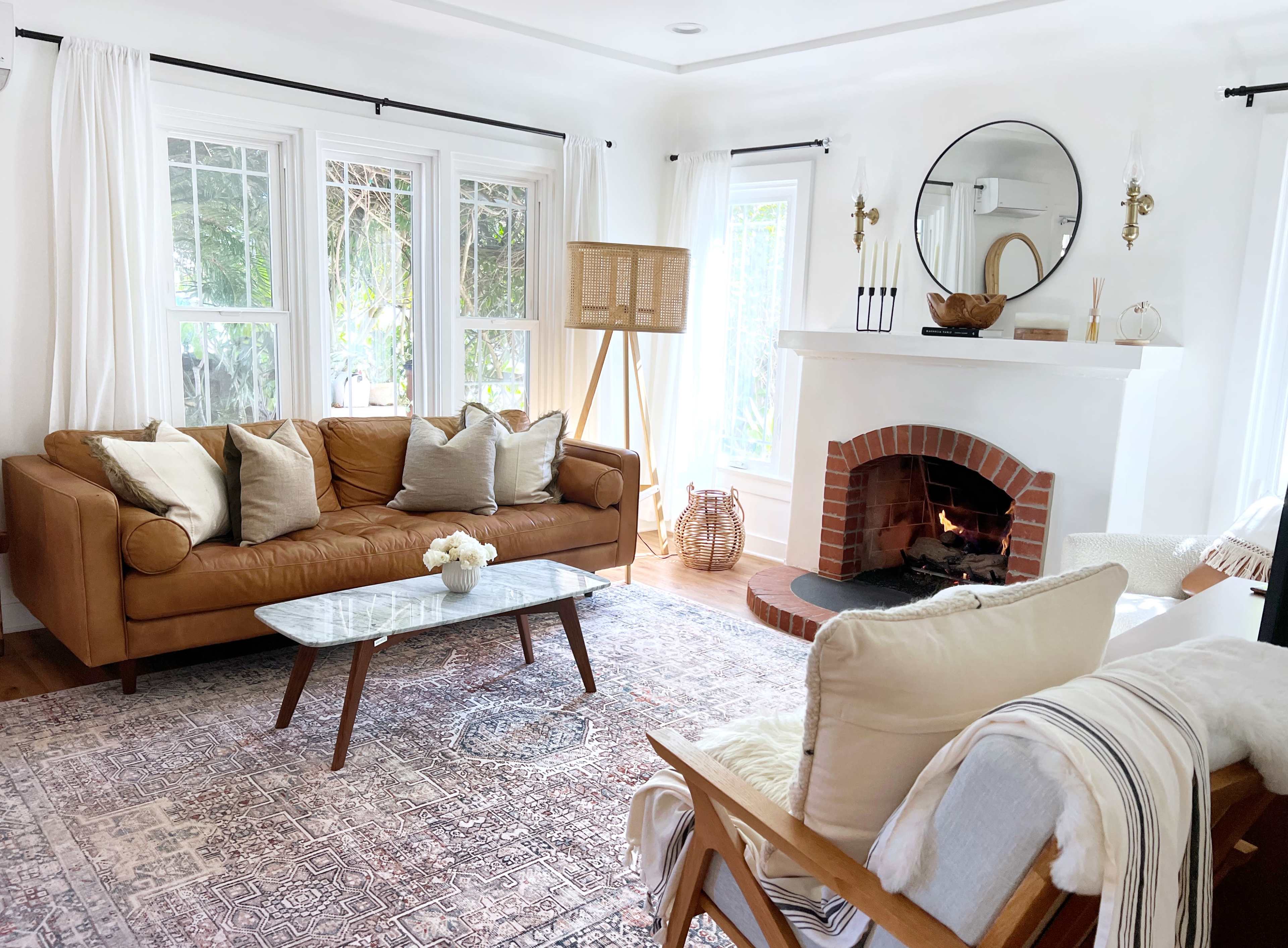 The living room features a brown leather sofa, a glass coffee table, and a fireplace with a circular mirror above it, surrounded by light curtains and natural decor elements.