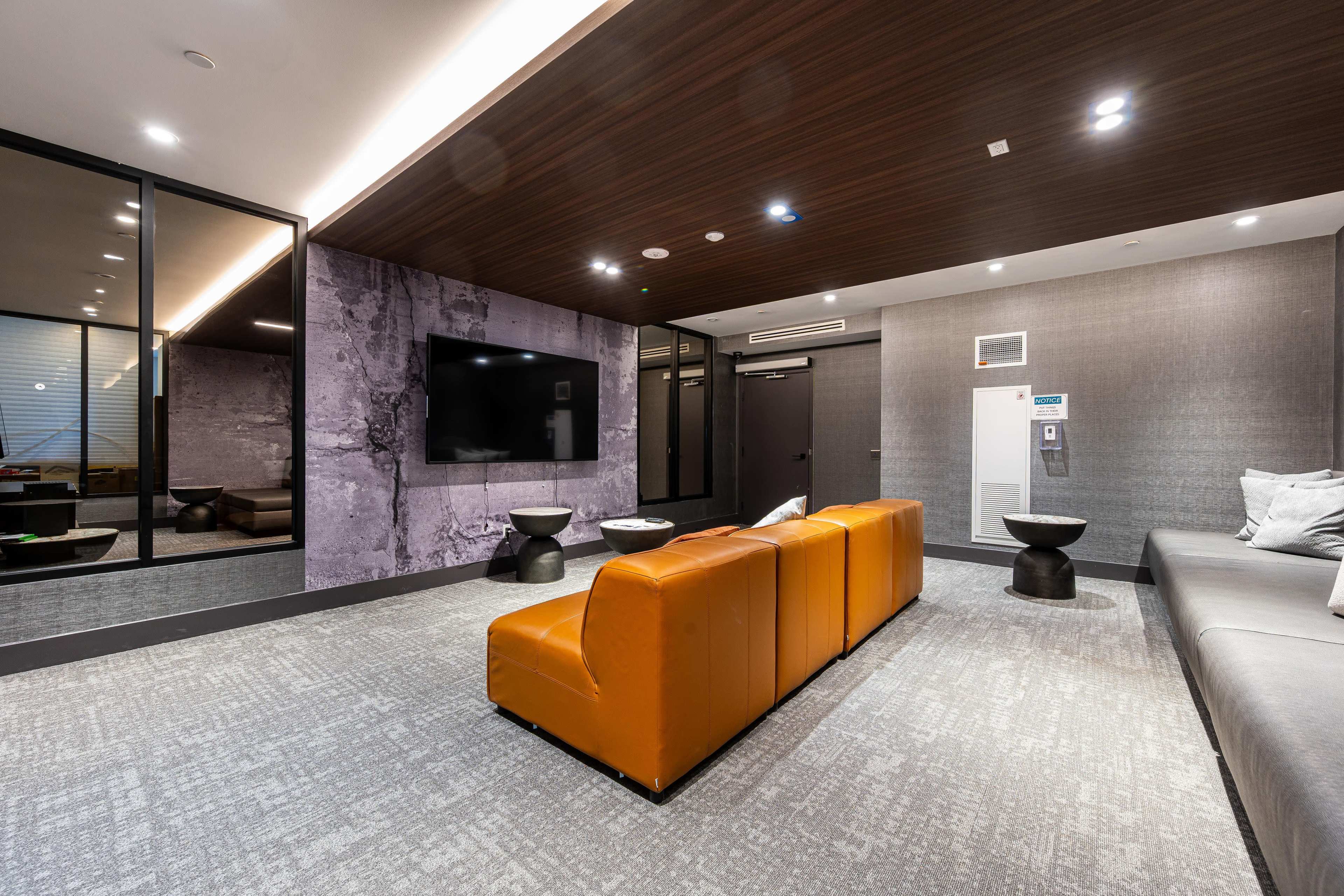 The image shows a modern lounge area with a large television, an orange leather couch, and a gray sofa, complemented by small black tables and a stylish wall design.