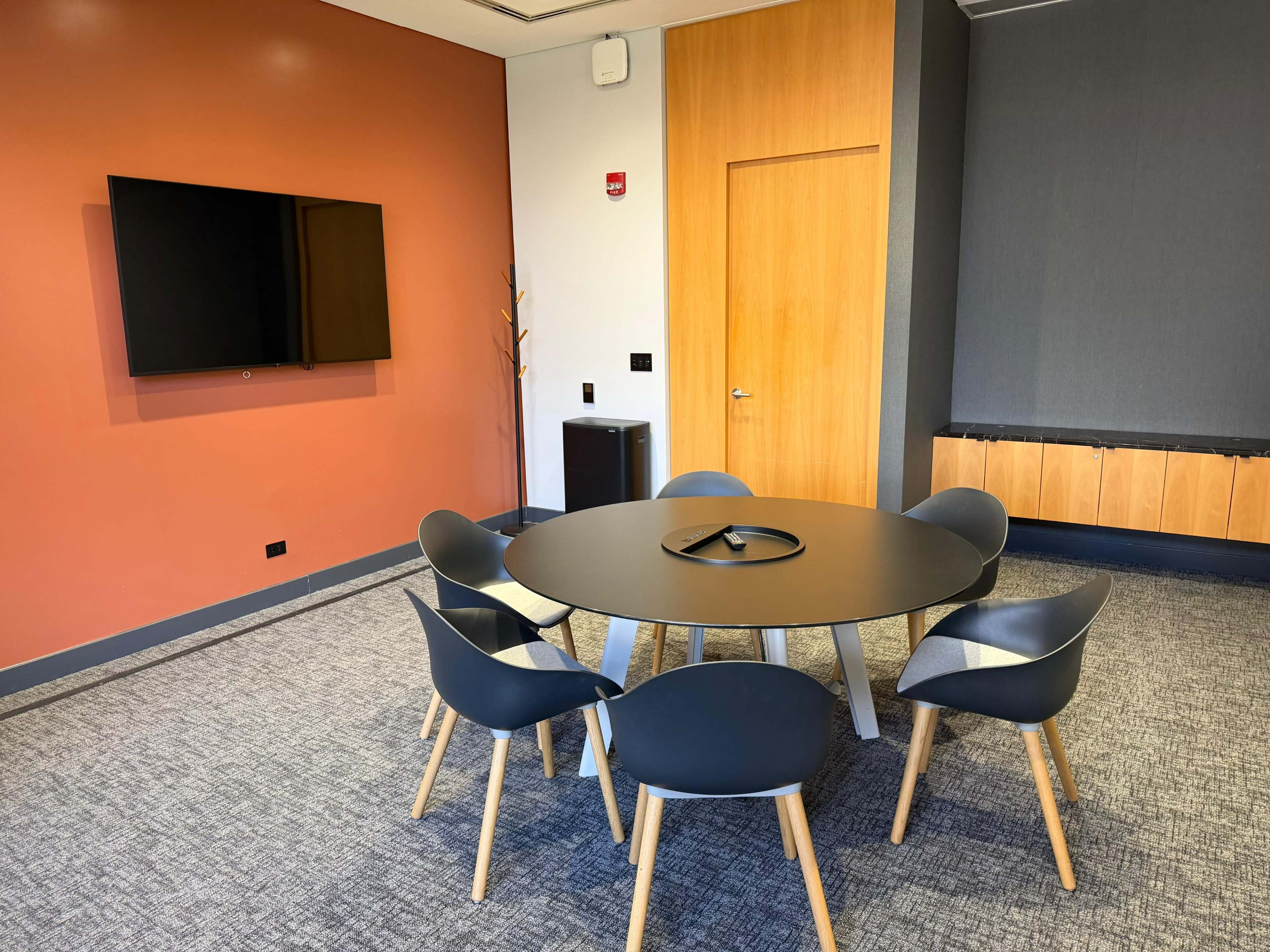 A conference room features a round table surrounded by six chairs, a mounted television on one wall, and a wooden accent along another wall.