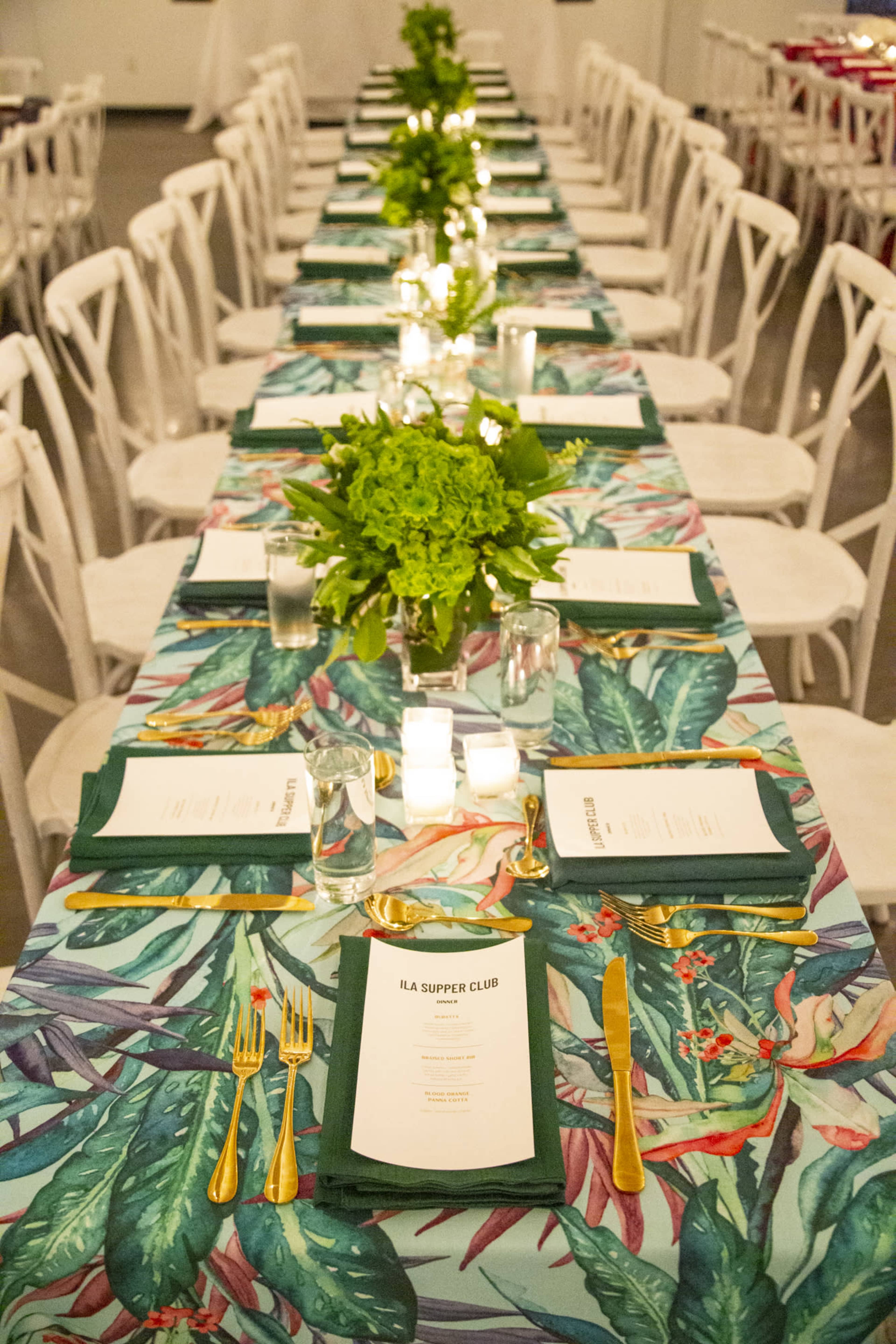 A long dining table is set with green menus, golden utensils, and candlelight, adorned with a tropical leaf-patterned tablecloth and greenery centerpieces.