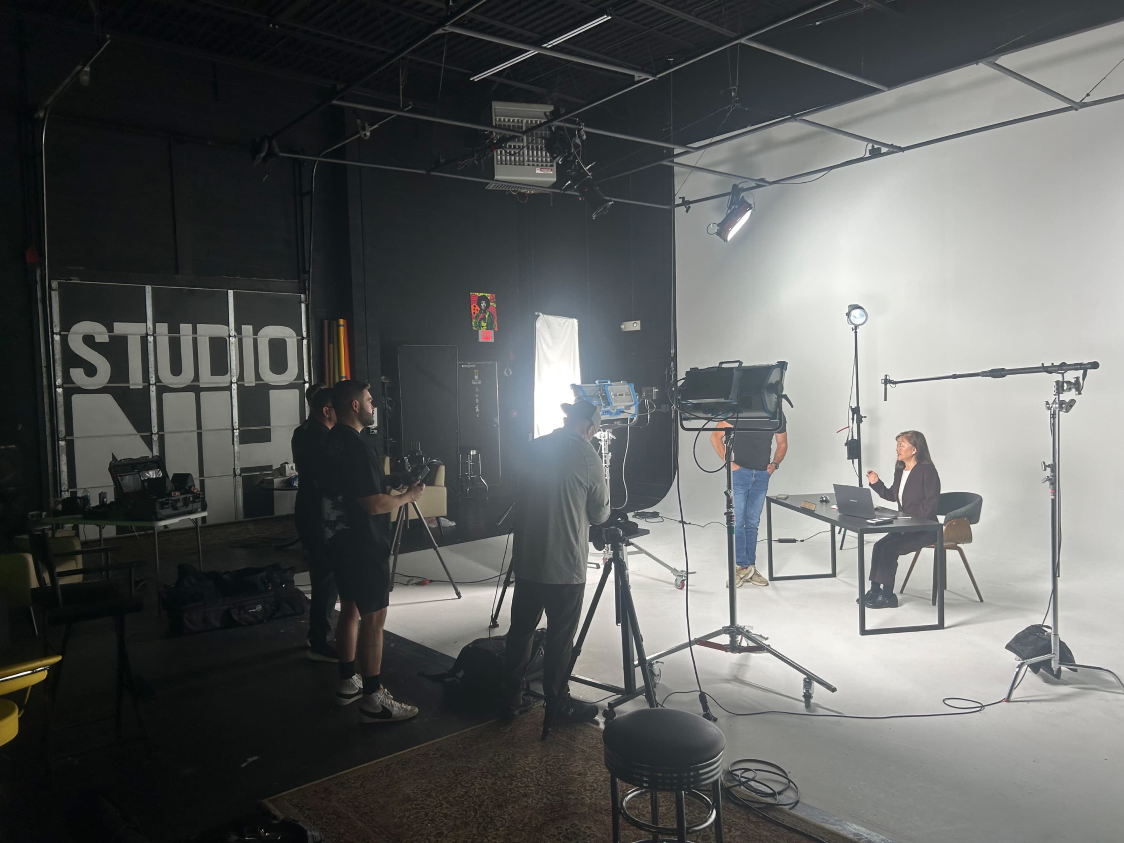 The image shows a film set in a studio with multiple crew members operating cameras and lights while a woman is seated at a table, facing a computer.