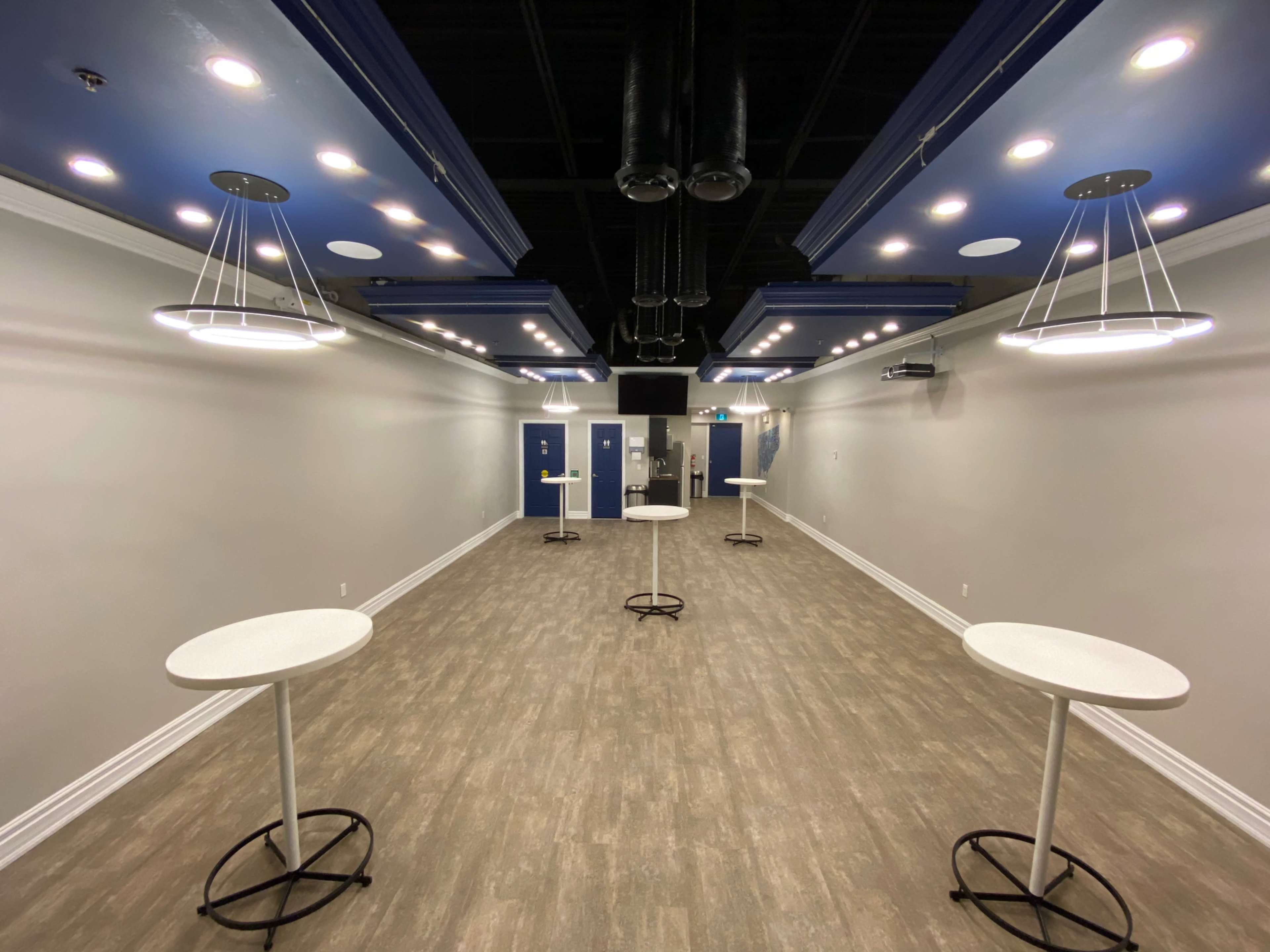 A modern indoor space with white tables, blue ceiling fixtures, and a gray wall, illuminated by overhead lights.