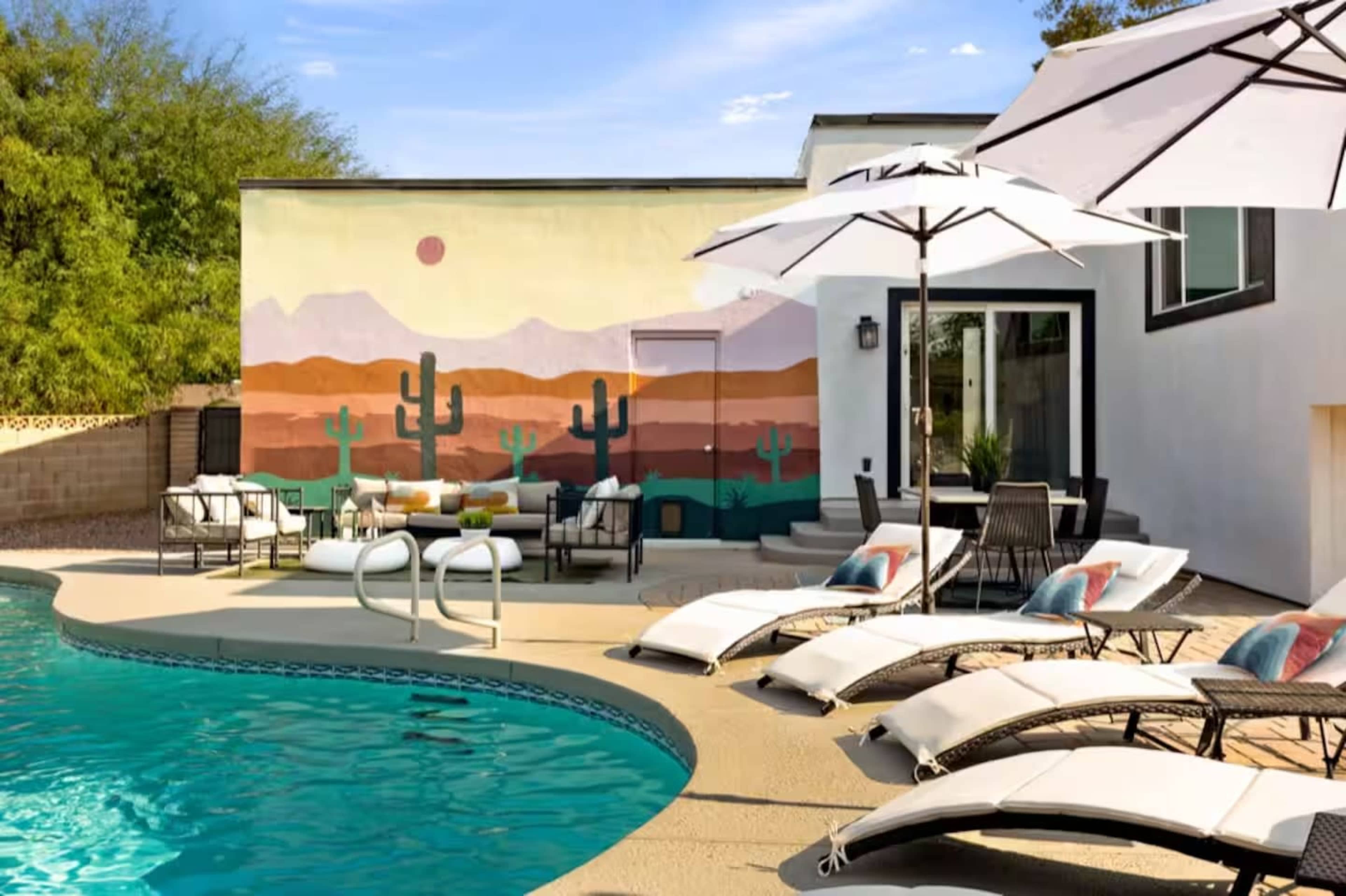 A backyard pool area features lounge chairs, large umbrellas, and a wall mural depicting a desert landscape with cacti and mountains.