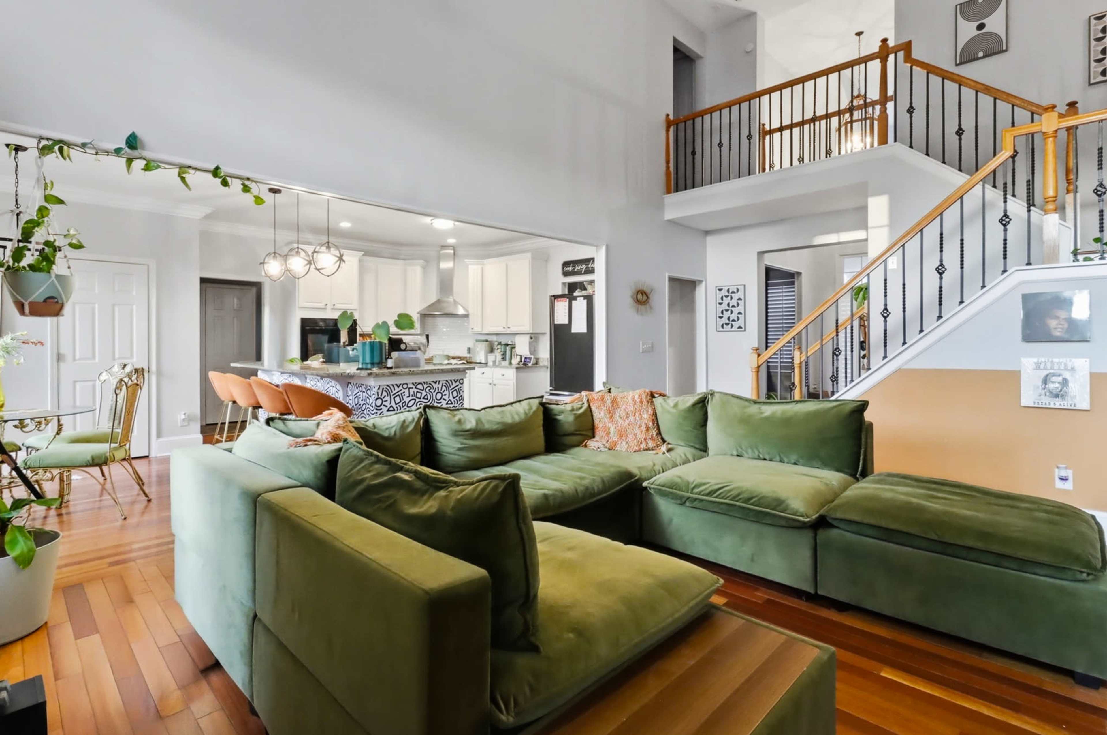 The image shows a spacious living area featuring a large green sectional sofa, a staircase with wooden railings, and a view of a modern kitchen in the background.