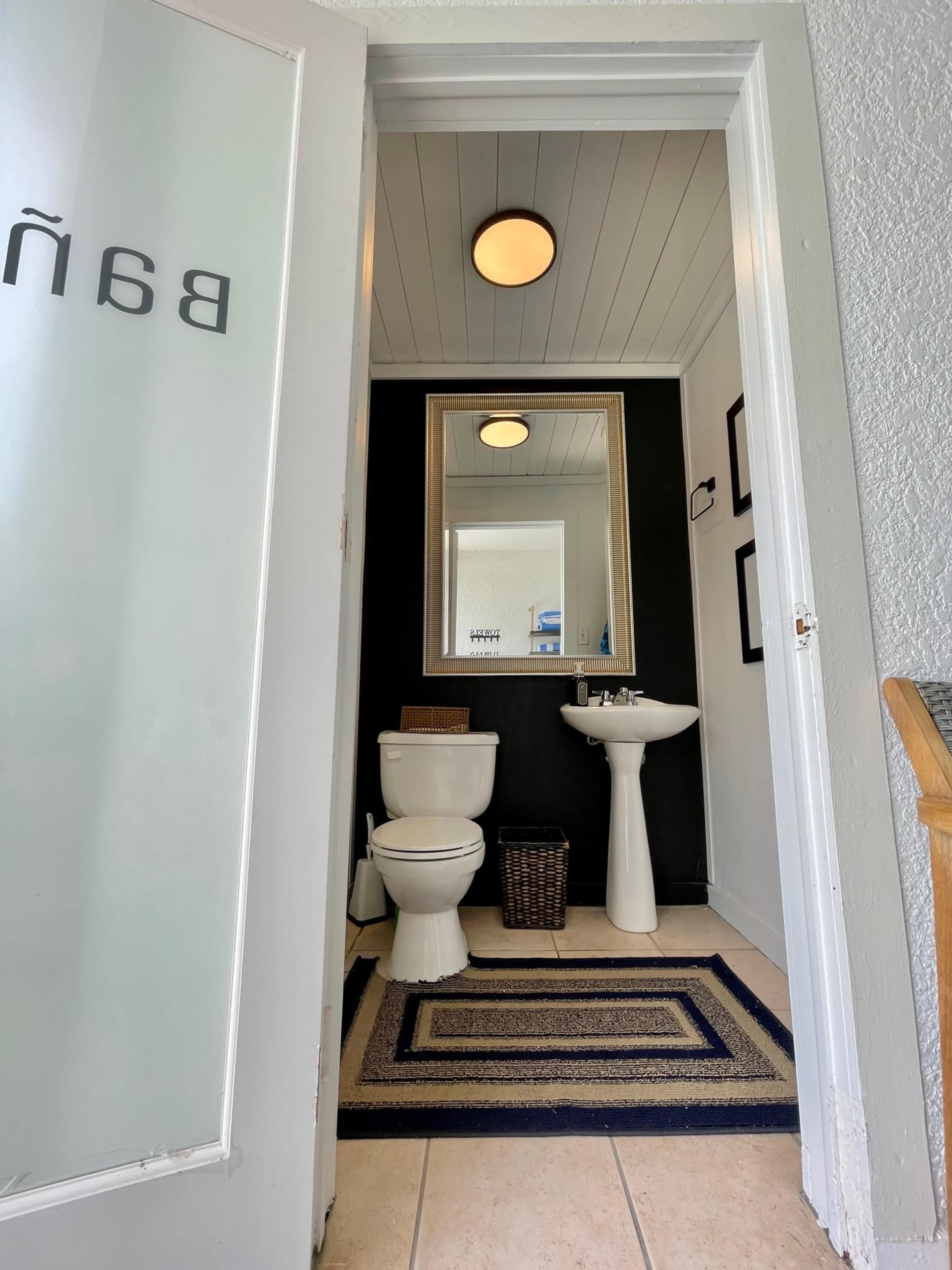 A clean bathroom entrance with a toilet, sink, and a large mirror, featuring a dark wall and a rug on the tiled floor.