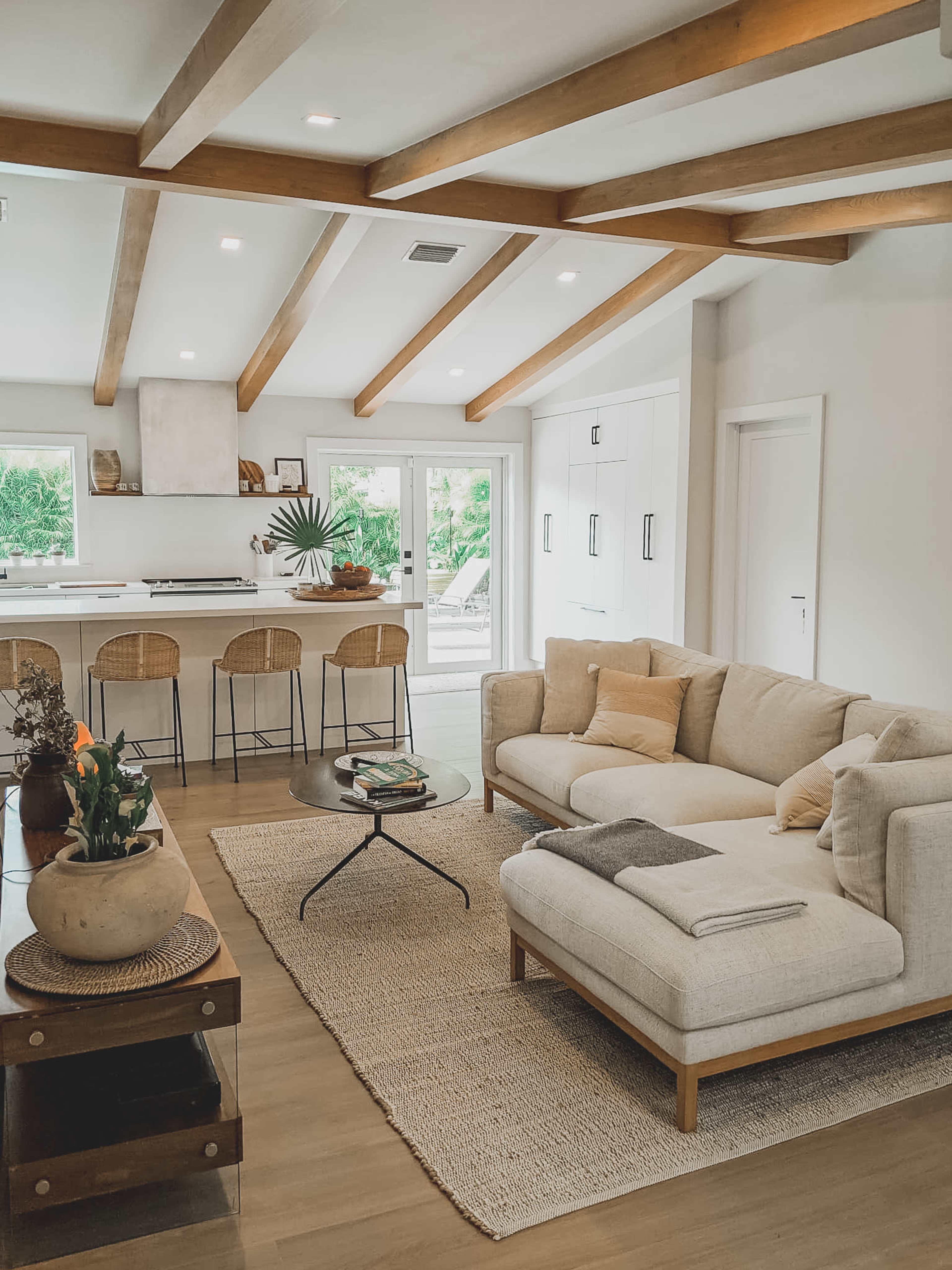 A modern living room features a light-colored sectional sofa, a round coffee table, and a kitchen area with bar stools, illuminated by natural light from sliding glass doors.