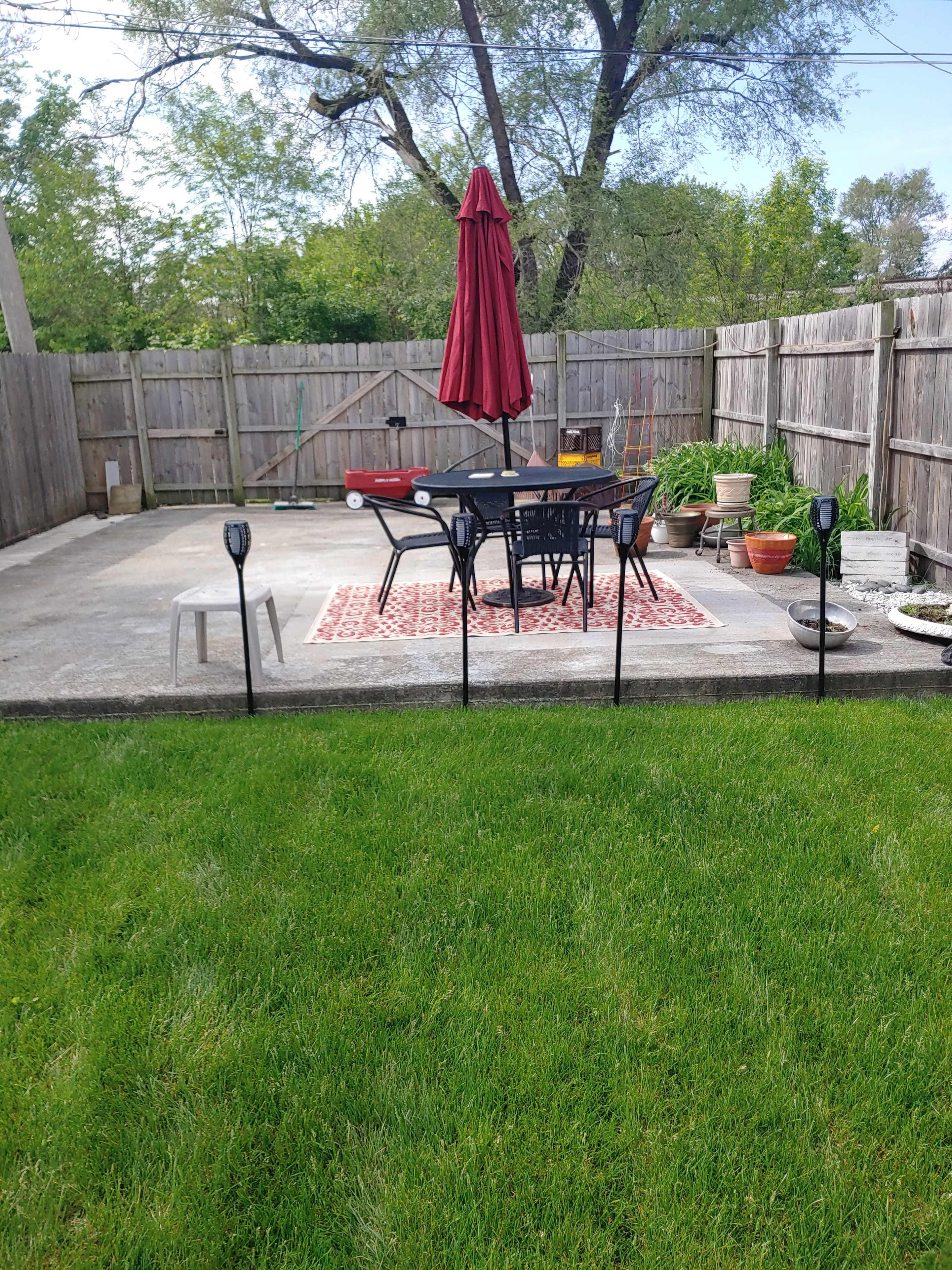 A patio area features a table and chairs under a red umbrella, surrounded by a lawn and bordered by wooden fencing.