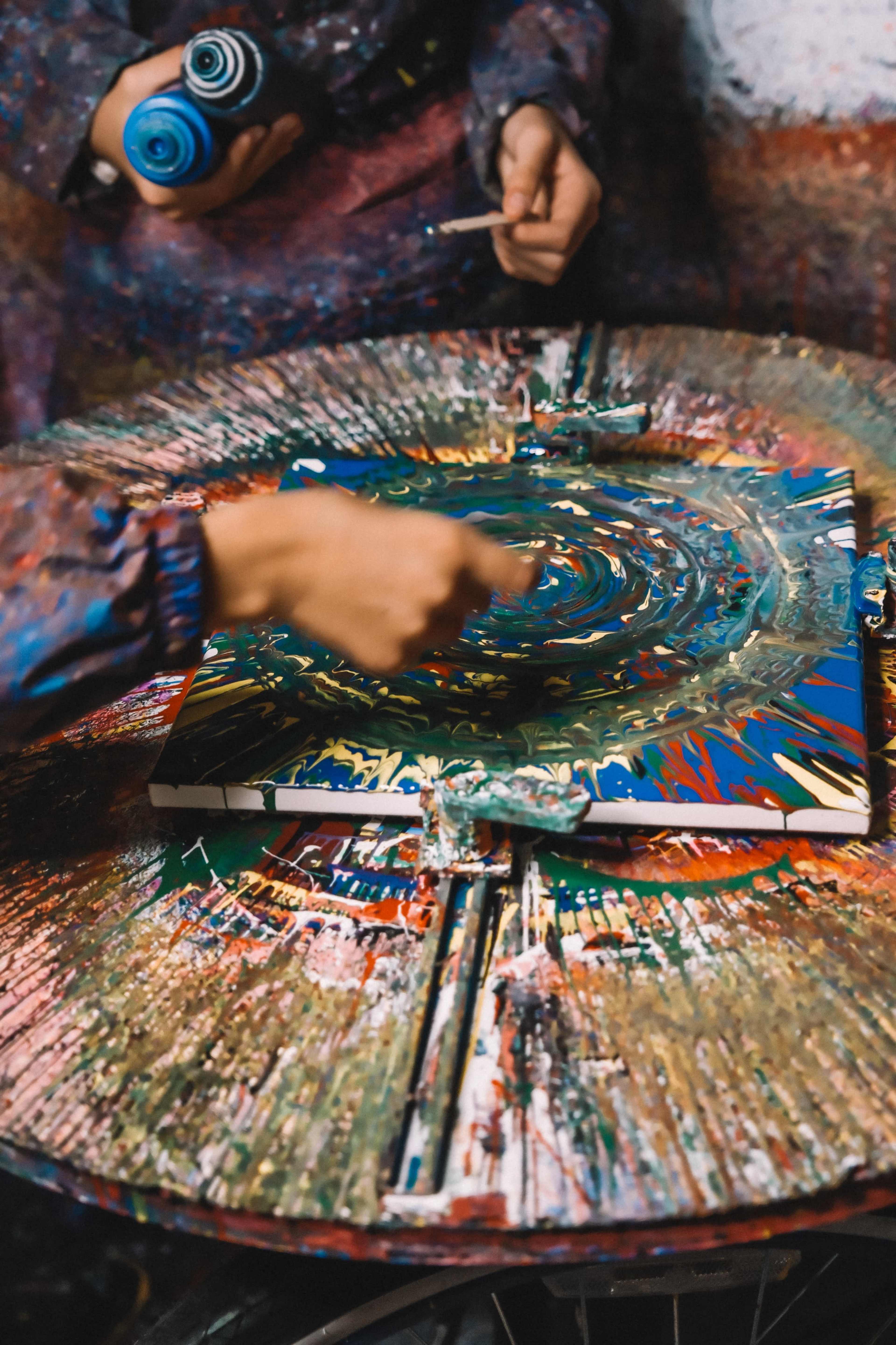 A person applies colorful paint to a canvas on a circular table covered in splattered paint.