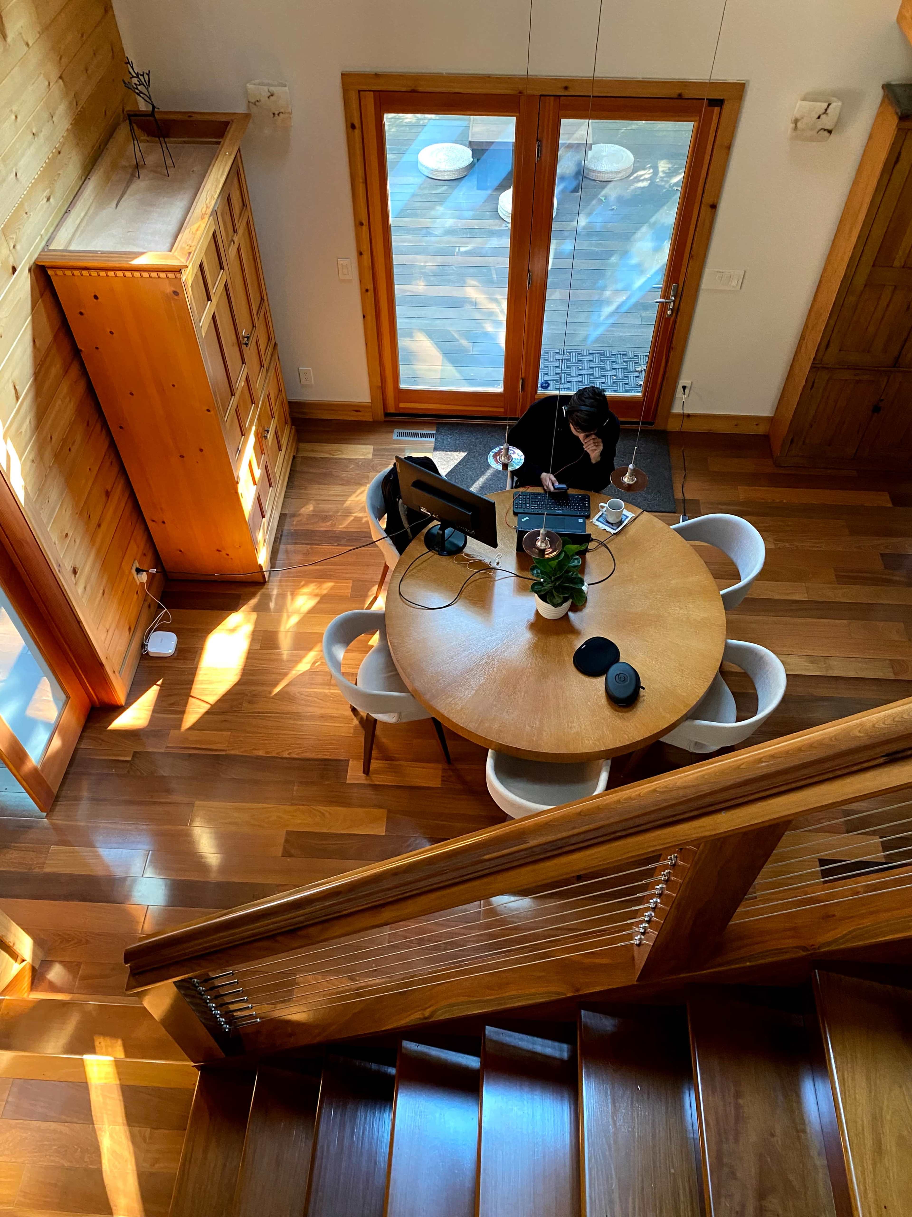 A person is working at a circular wooden table in a well-lit room with large glass doors and wooden flooring.