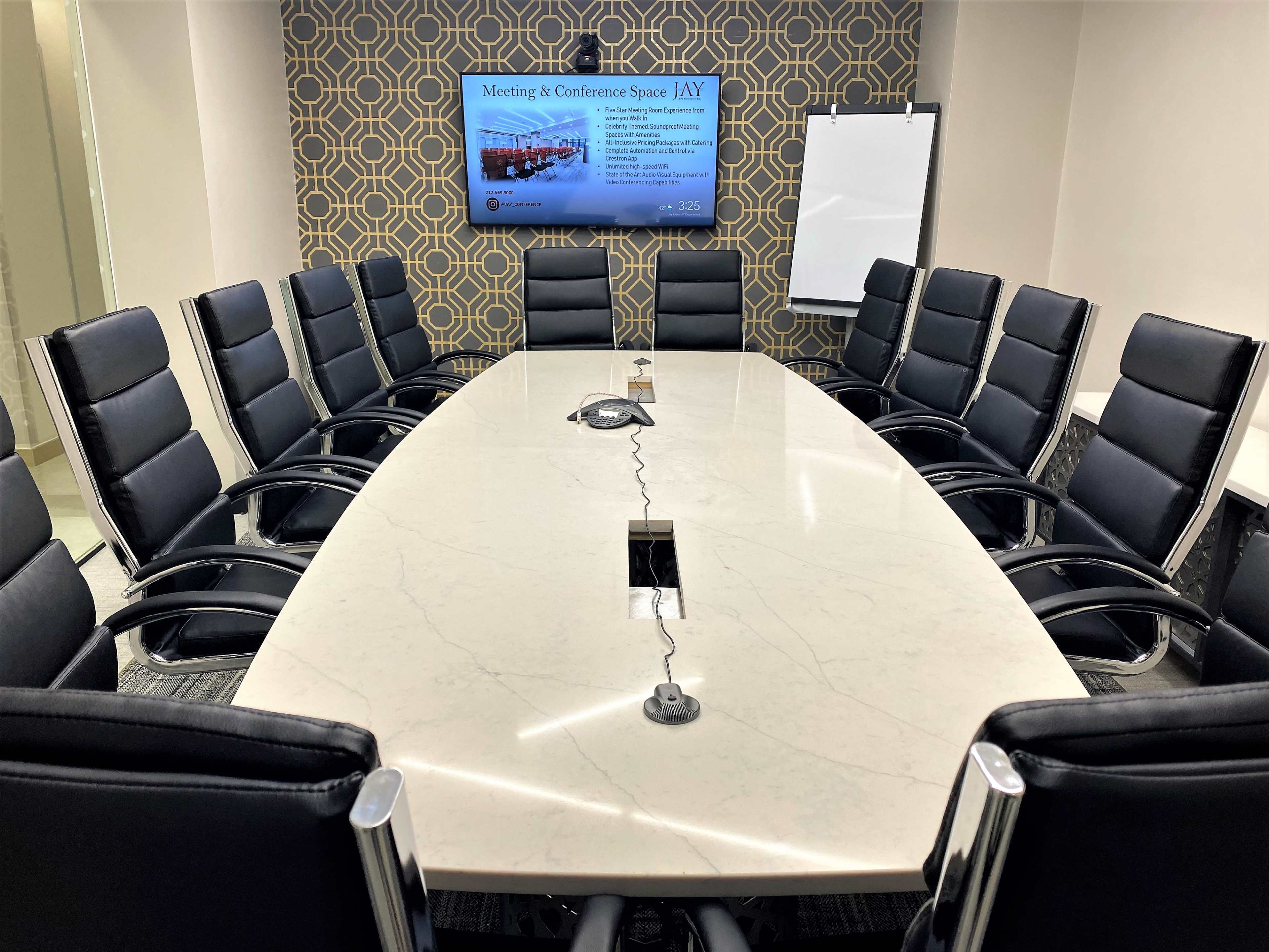 A large conference room features a long oval table surrounded by black leather chairs, with a wall-mounted screen displaying information and a whiteboard at the side.