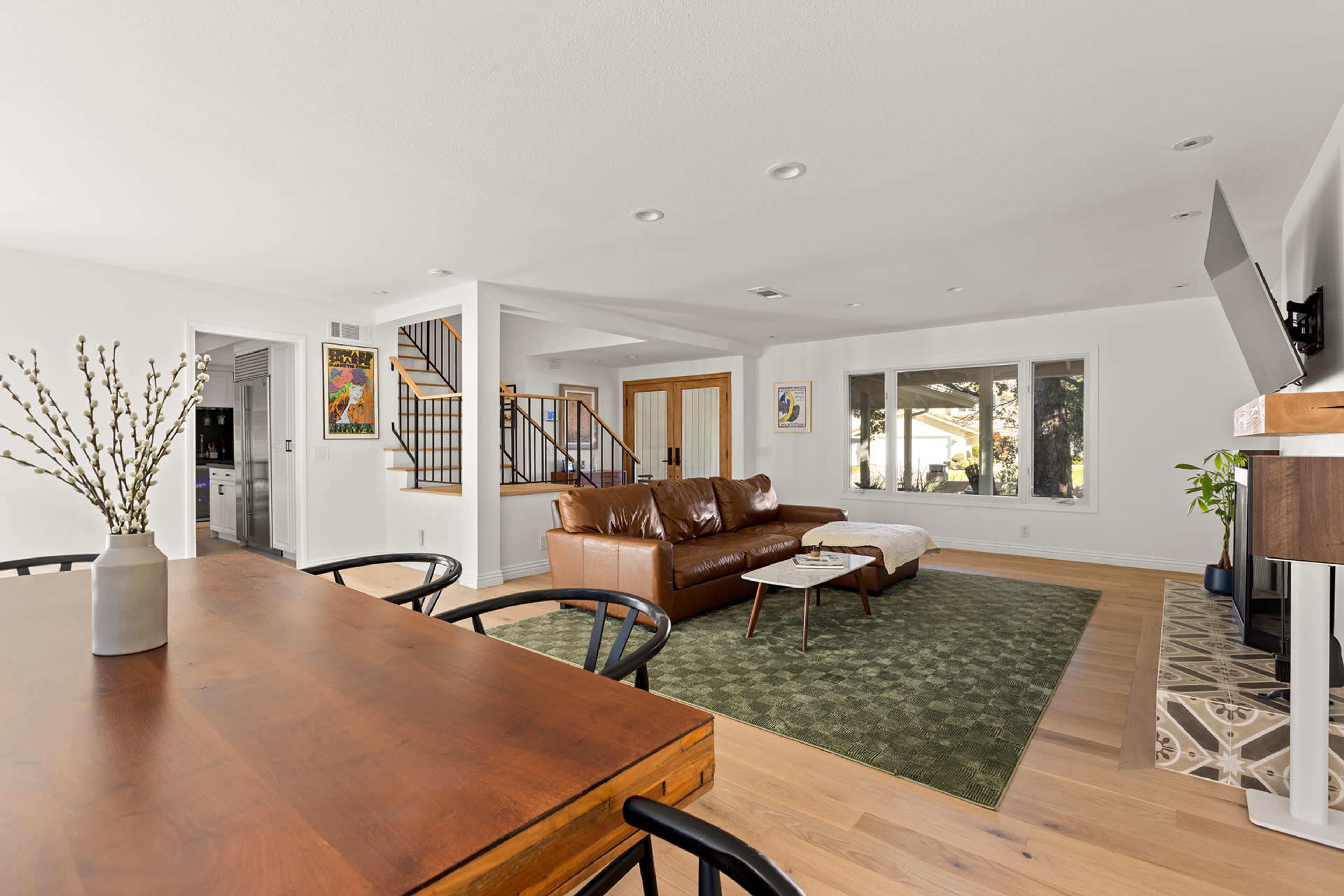 A spacious, modern living area featuring a brown leather sofa, a green rug, a wooden dining table, and large windows overlooking a patio.