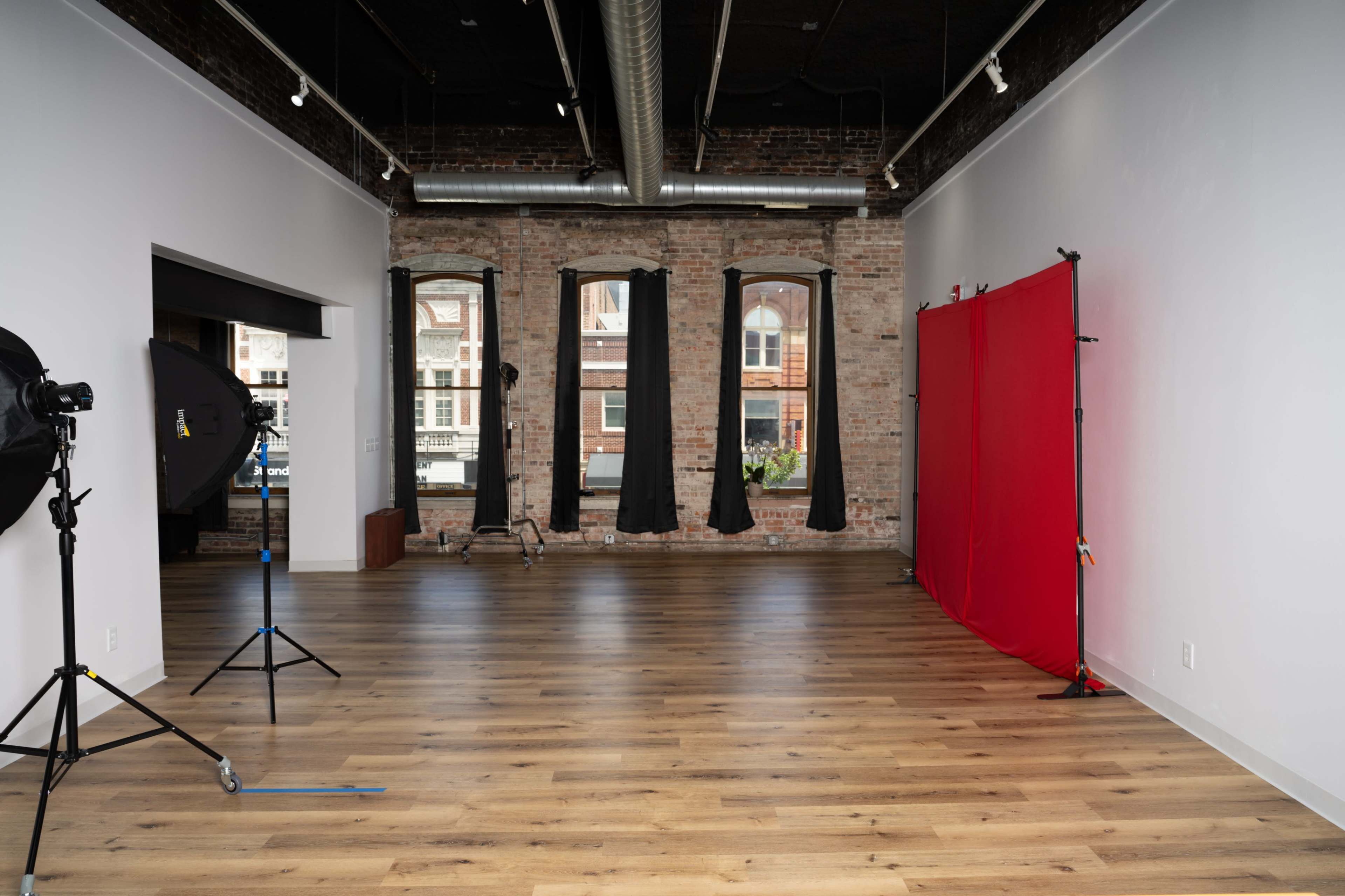 A spacious, empty photography studio with large windows, a red backdrop, and lighting equipment set up in the room.