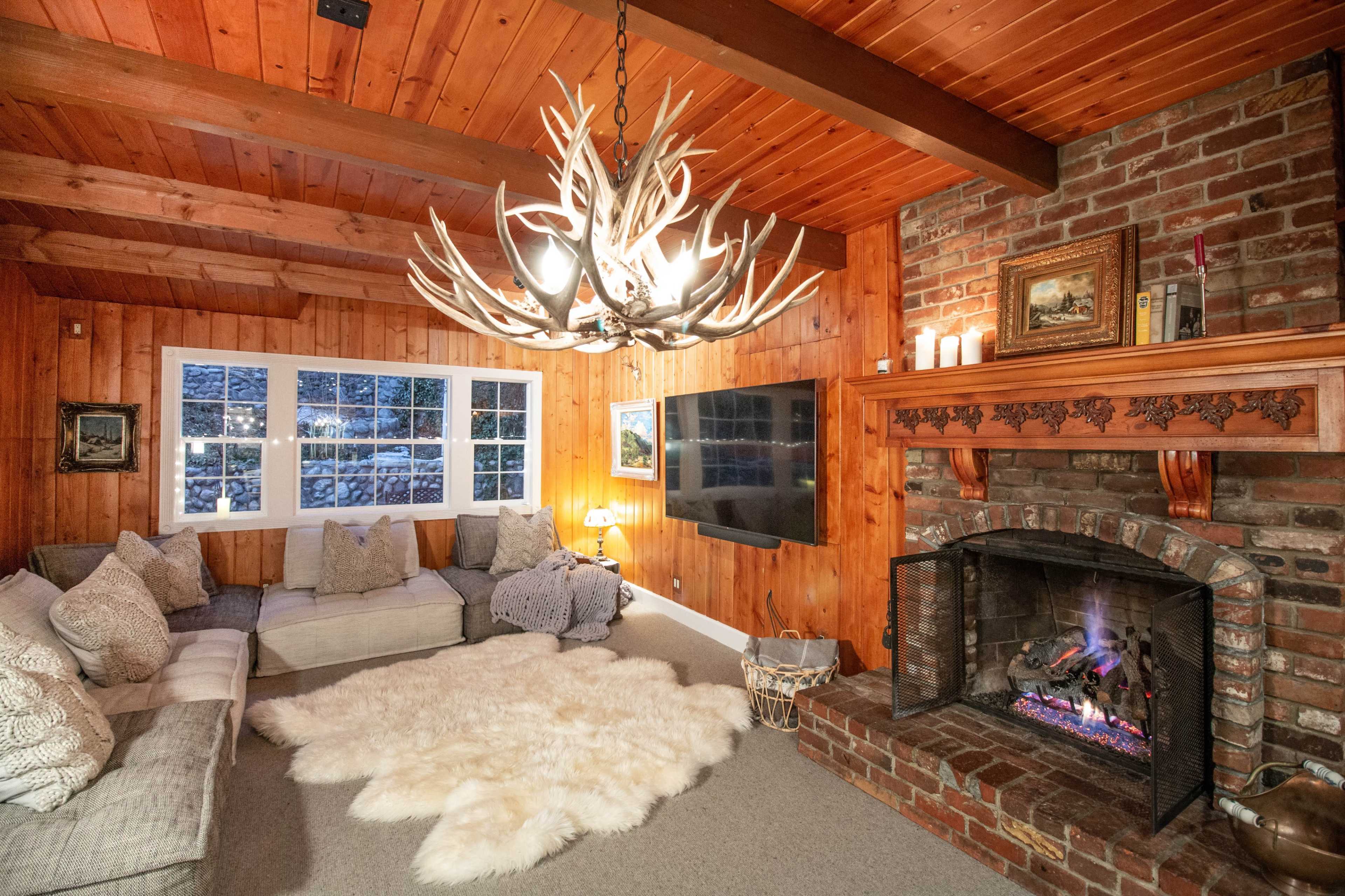 A cozy living room with wooden walls, a brick fireplace, a large antler chandelier, and a plush area rug in front of a sectional sofa.