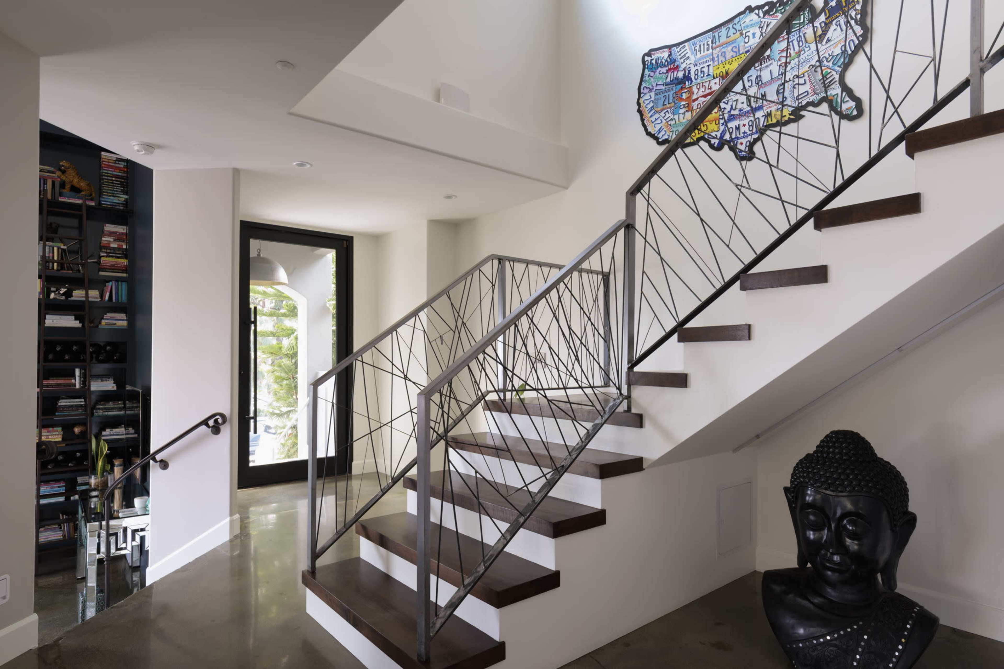 A modern staircase with a metal railing features a wooden handrail, and a black Buddha statue stands near the base, with a colorful art piece of the United States mounted on the wall above.